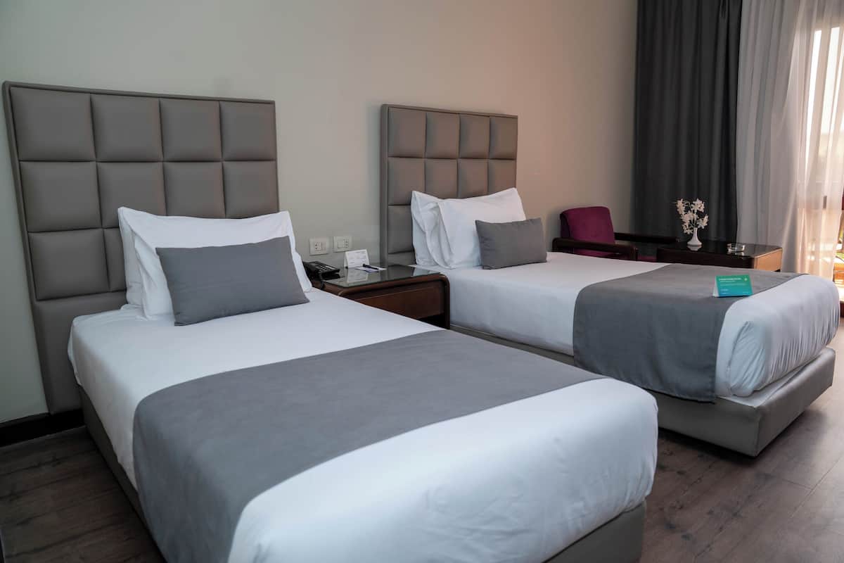 A hotel room with two beds, each with a padded headboard and white bedding. There is a desk with a chair and a vase with flowers on it.