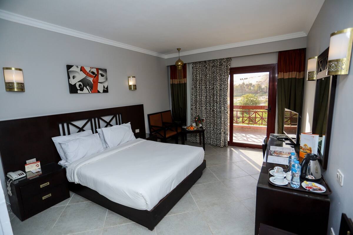 A hotel room with a large bed, a desk with a chair, a small table with a water bottle and a kettle, and a view of the outdoors through a sliding door.