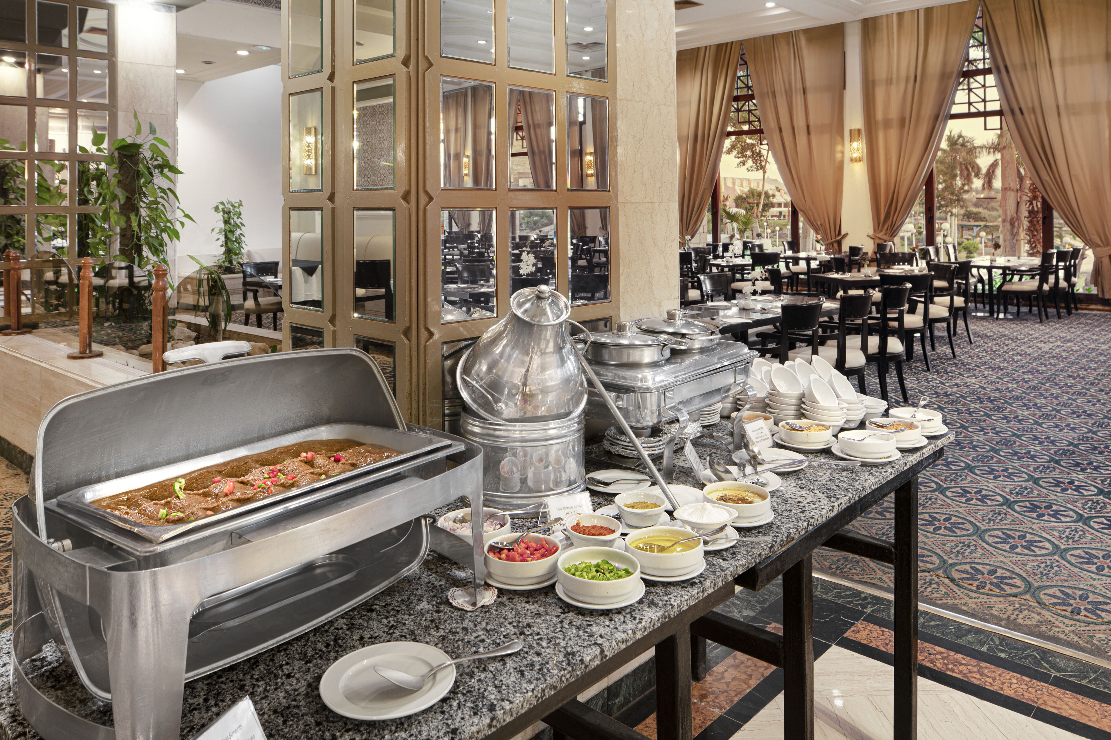 A buffet setup with various food items and a beverage station in a spacious dining area with a patterned floor and large windows.