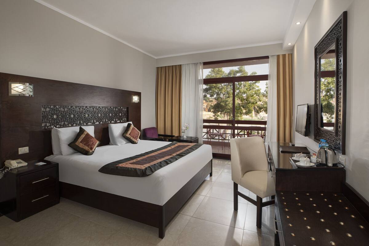 A hotel room with a large bed, a desk with a chair, a TV, and a balcony with a view of trees.