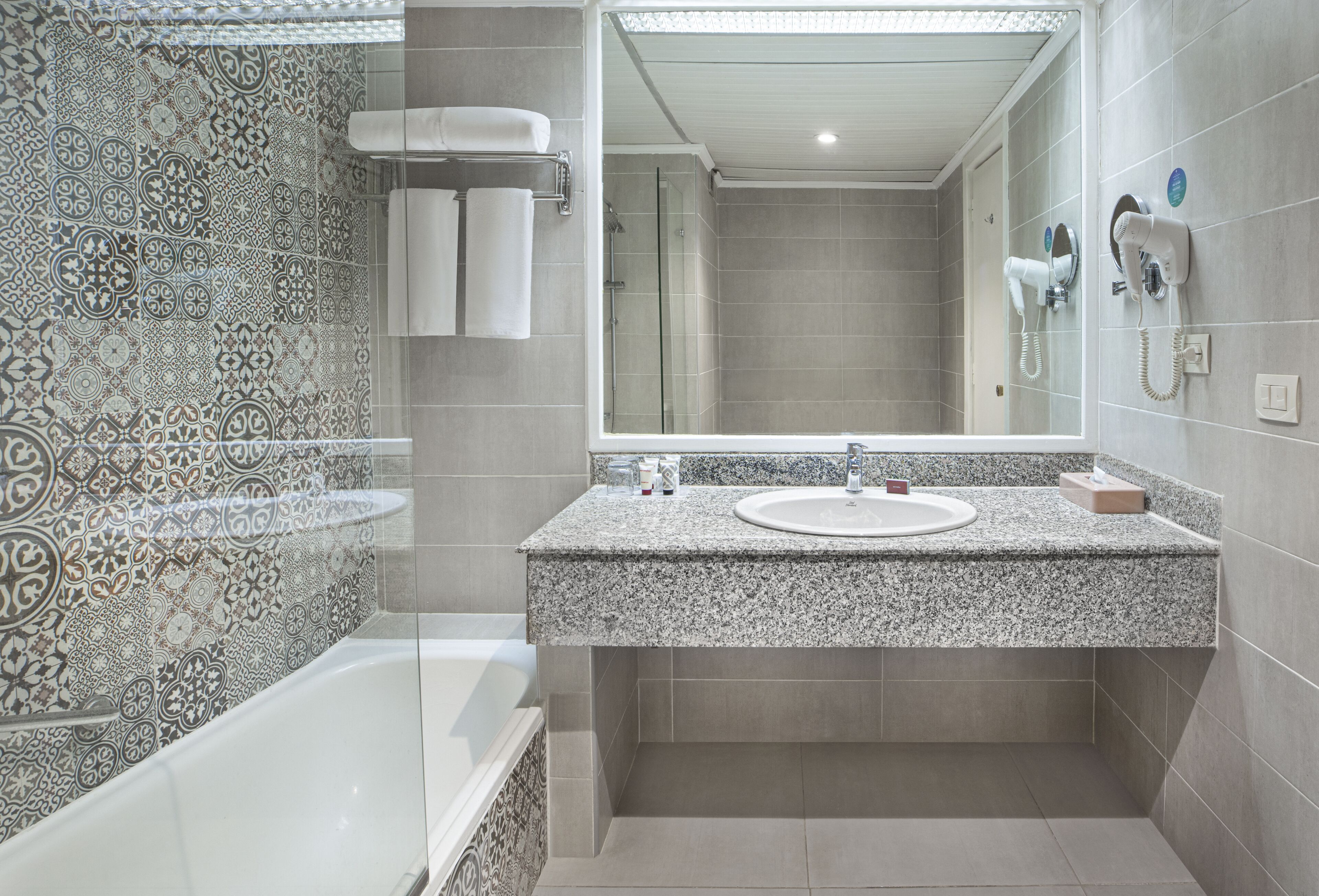 A bathroom with a patterned tile wall, a white bathtub, a granite countertop sink, and a large mirror.