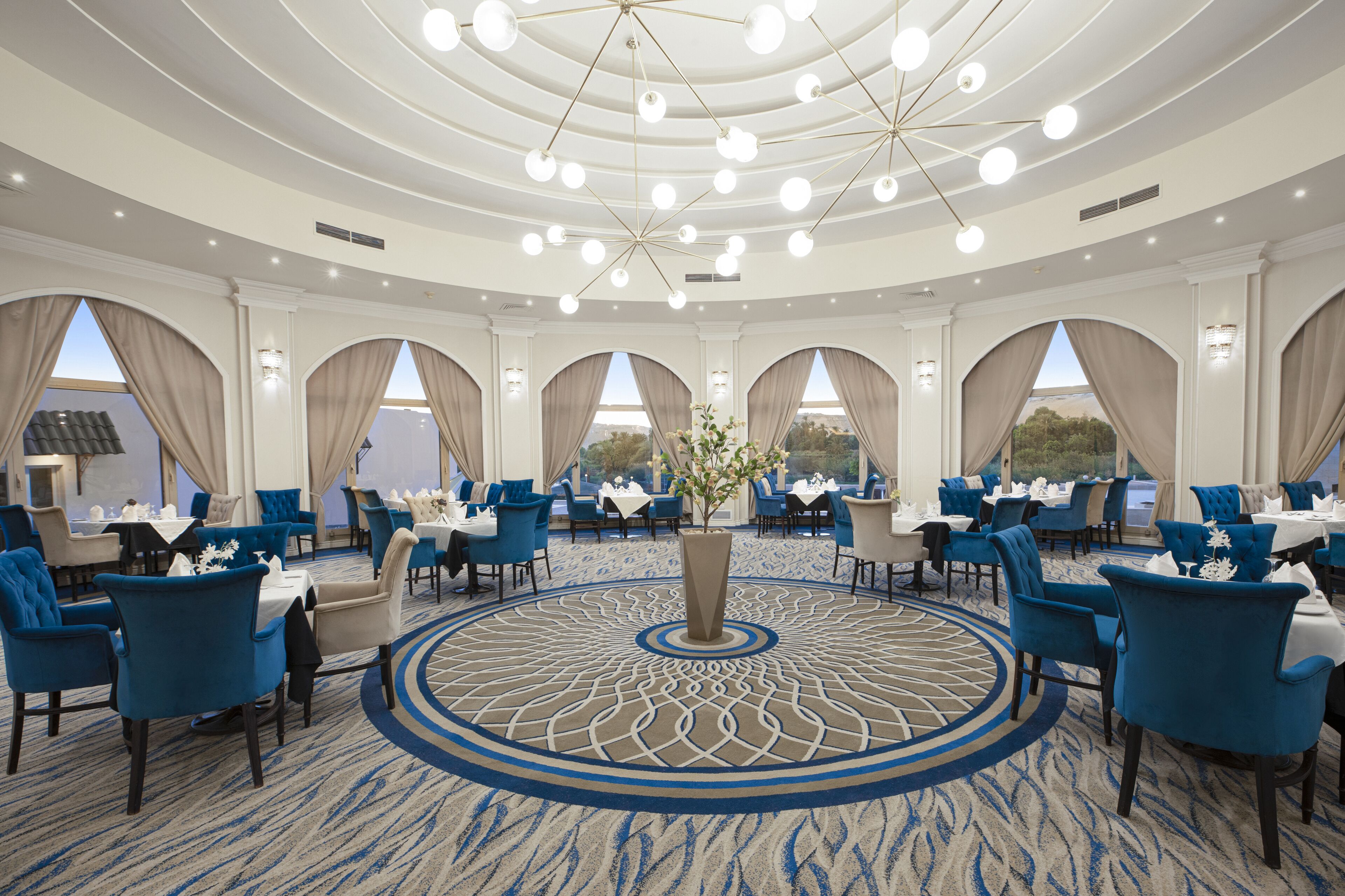 A spacious dining area with round tables and blue chairs, a decorative central rug, and large windows with curtains.