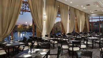 A spacious restaurant with multiple tables and chairs, large windows with curtains, and a view of a body of water.