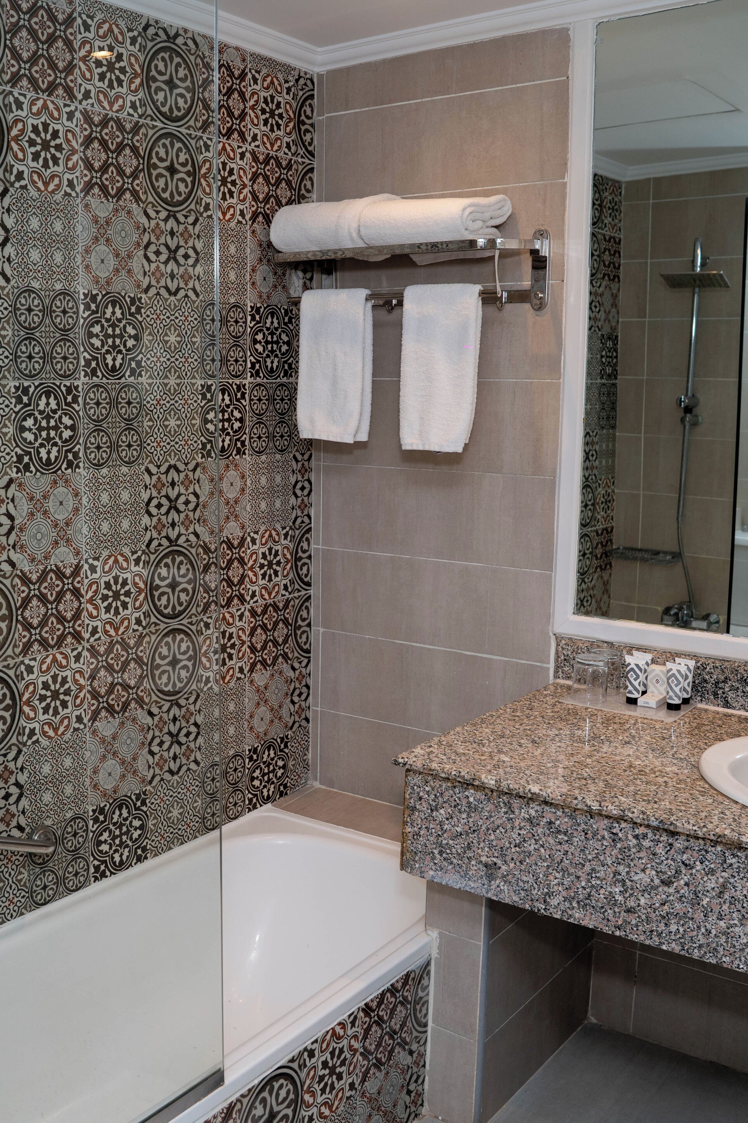 A bathroom with patterned tiles, a white bathtub, a sink with a granite countertop, and a mirror.