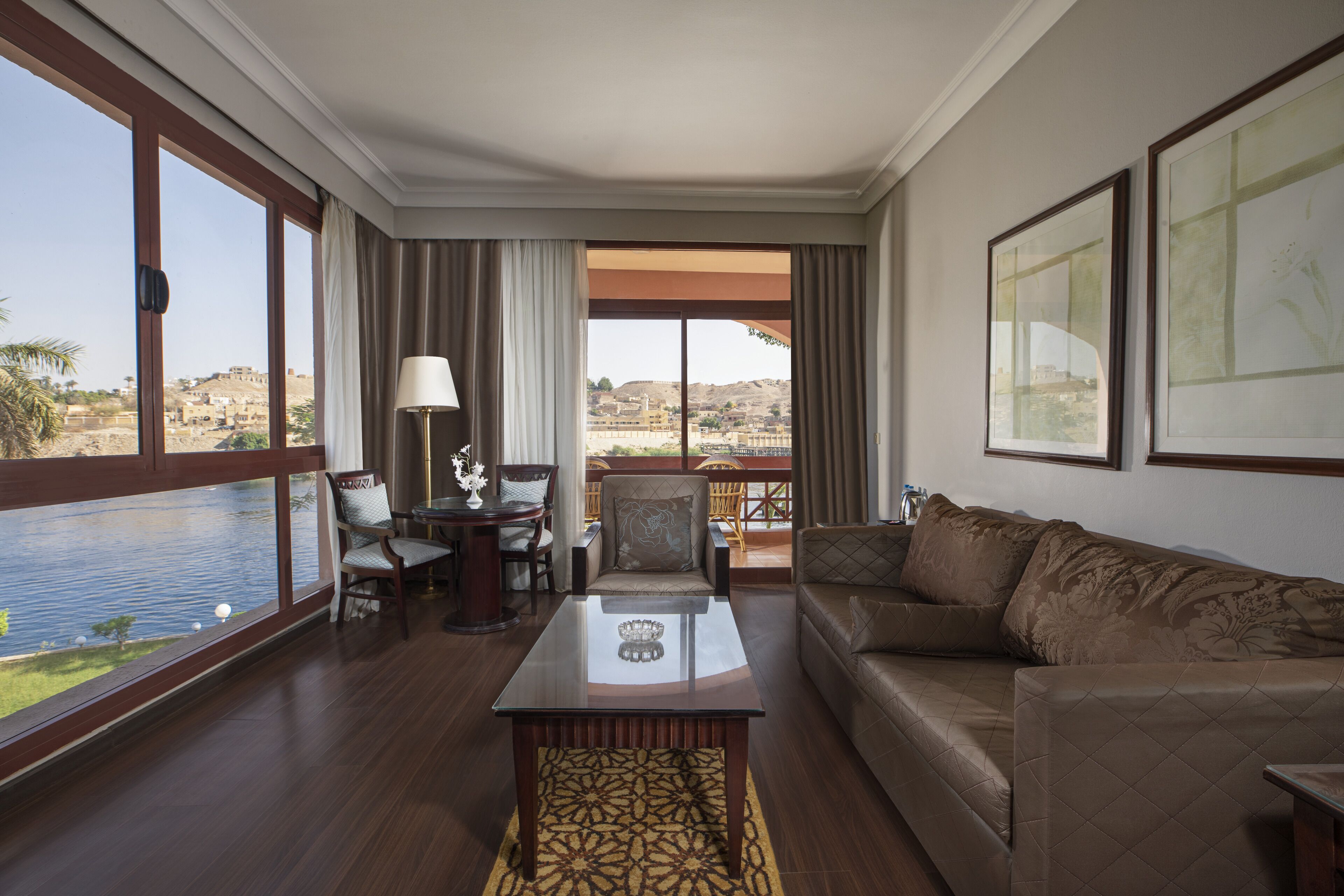 A spacious living room with a large window offering a view of a body of water and distant hills. There is a leather sofa, a glass coffee table, and a dining area with chairs and a table.