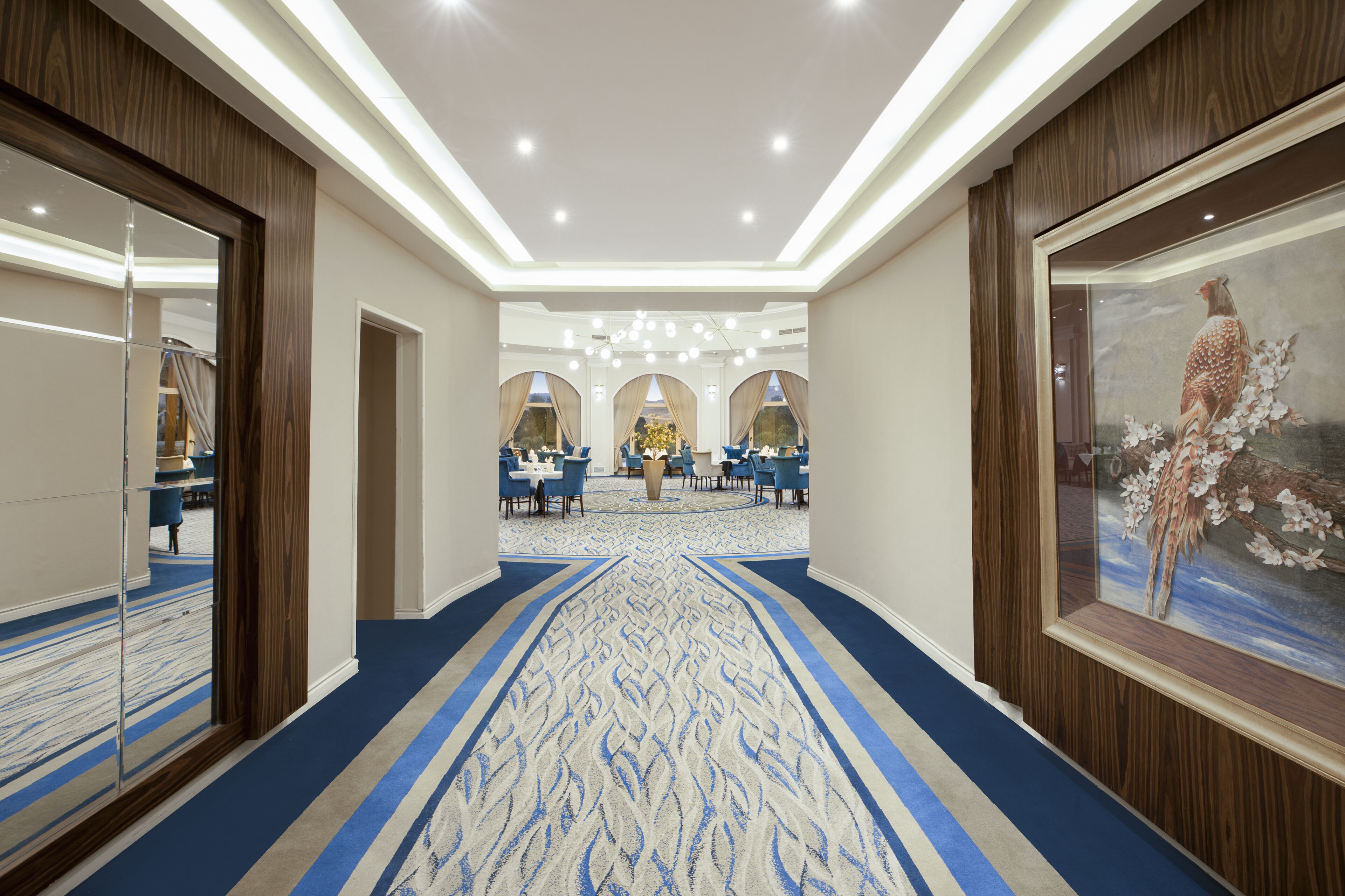 Un pasillo de hotel con alfombra estampada, un área de comedor con mesas y sillas, y una obra de arte enmarcada en la pared.