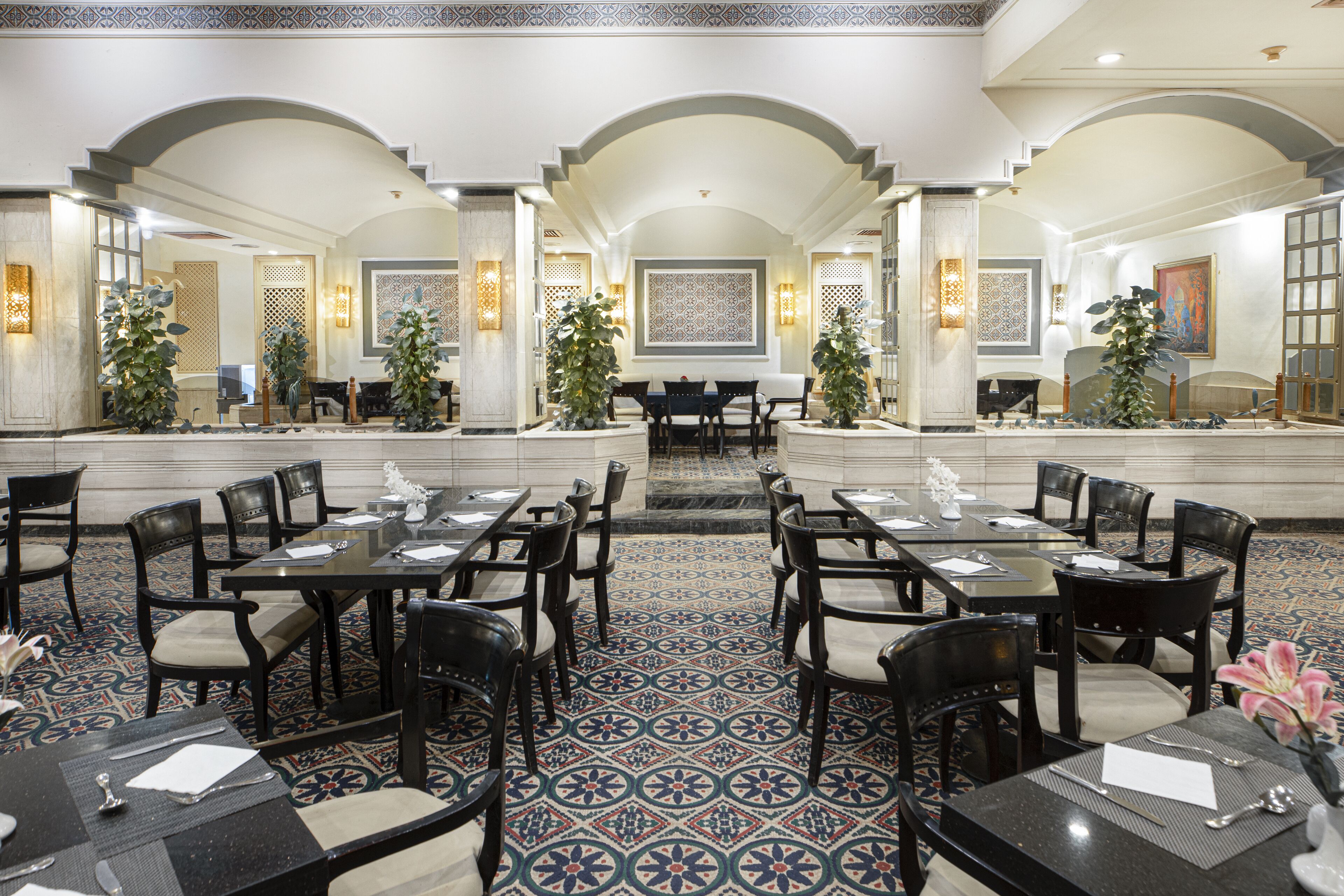 A hotel lobby with a central staircase, multiple dining tables with chairs, and decorative plants.