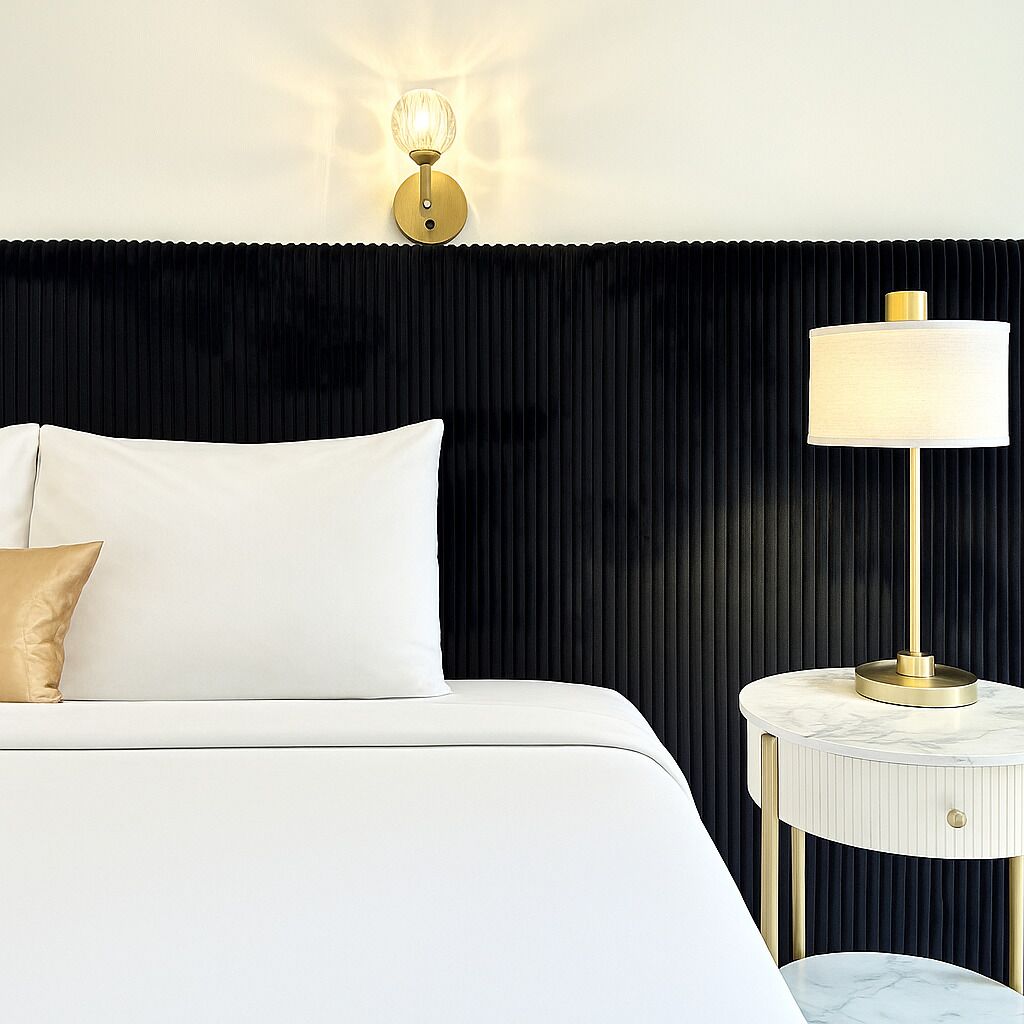 Una cama bien tendida con sábanas blancas y una sola almohada color beige, una mesita de noche redonda con una encimera de mármol blanco y una lámpara de pared con base dorada y pantalla blanca.