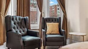 Quarto com duas poltronas de couro preto, uma mesa redonda pequena e vista para edifícios através de uma janela com cortinas.