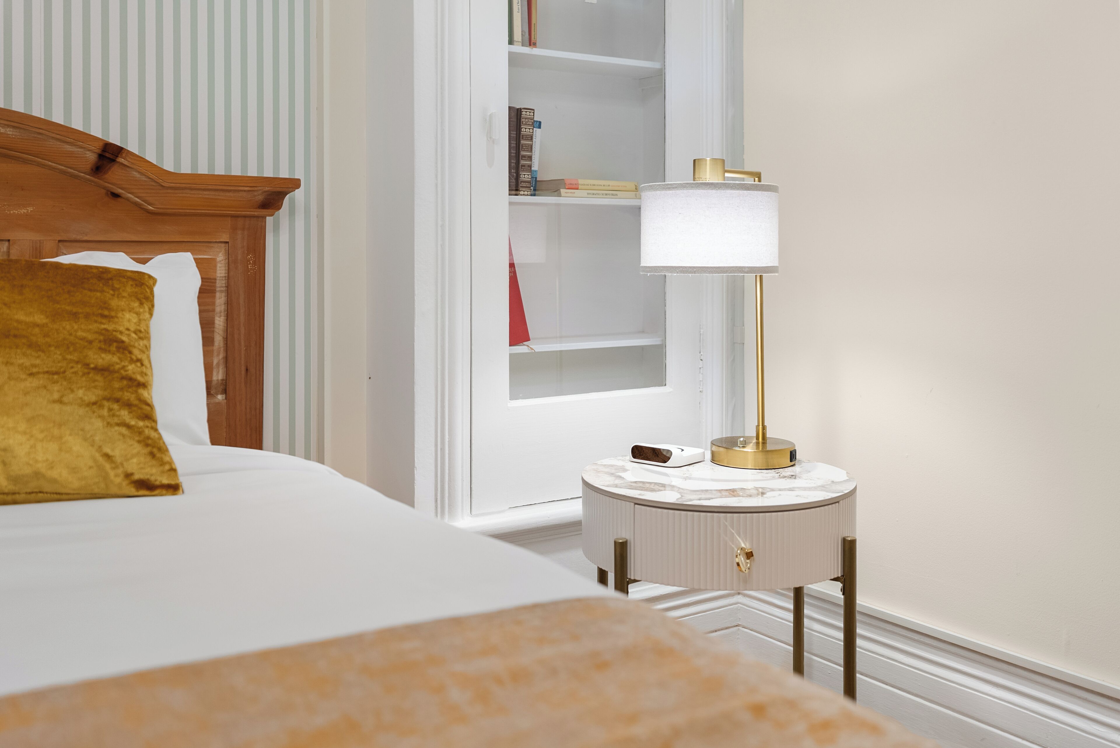 Habitación de hotel ordenada con cama, mesita de noche con lámpara y estantería.