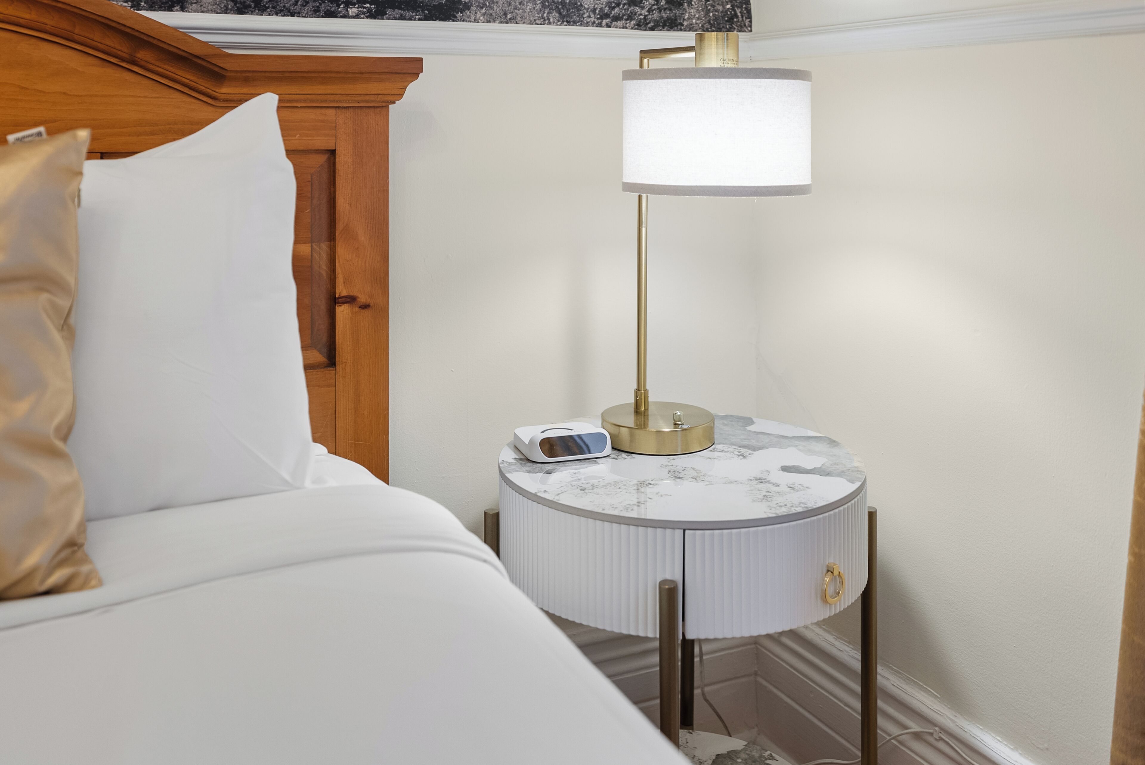 Una cama bien hecha con sábanas blancas y un almohadón color beige, una mesita redonda con cubierta de mármol, una lámpara moderna y un pequeño dispositivo electrónico.