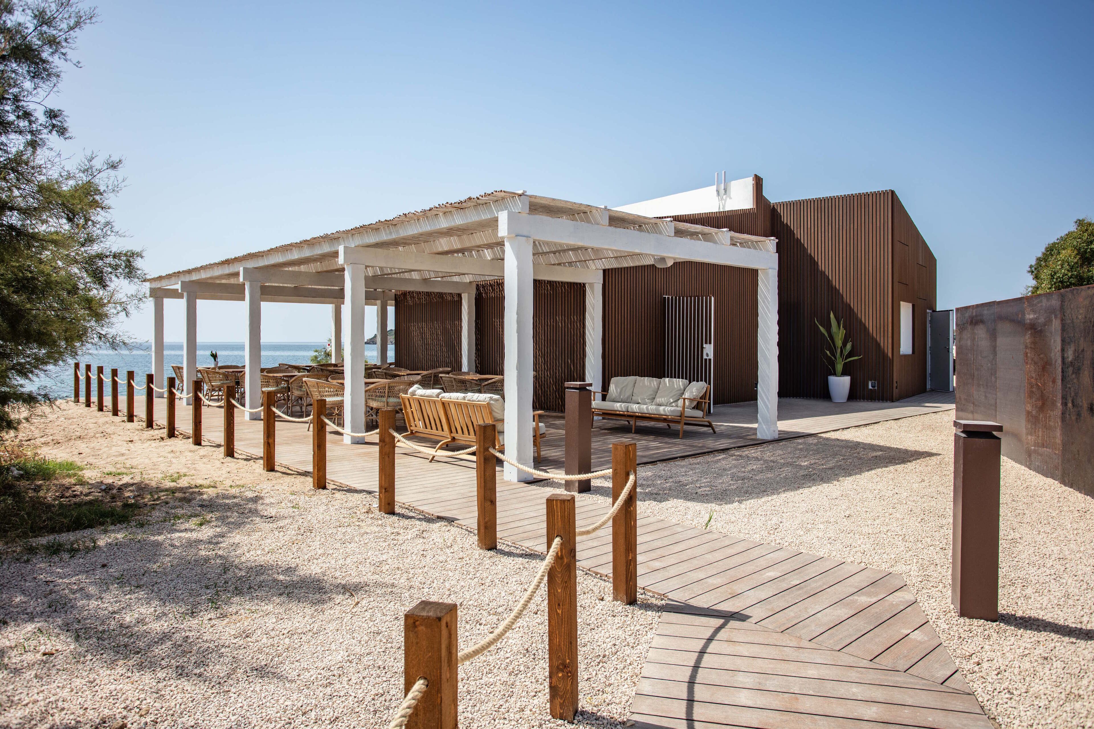 A beachside outdoor seating area with wooden tables and chairs, a covered patio, and a view of the sea.