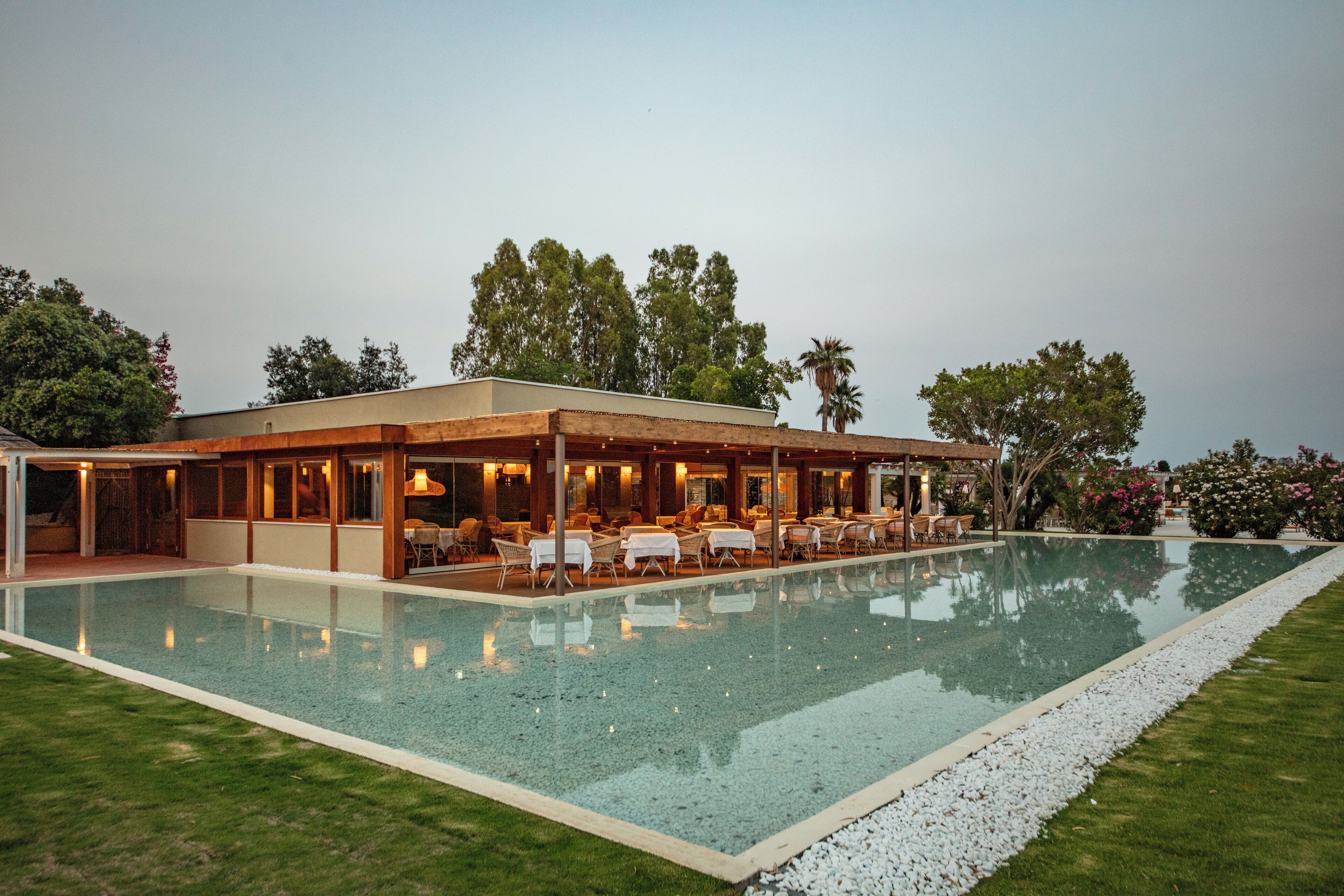 Un área de piscina moderna al aire libre con sillones de descanso y una zona de estar cubierta, rodeada de follaje y árboles.