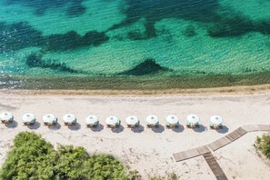 An aerial view of a beach with a row of beach umbrellas and lounge chairs, a wooden walkway, and clear turquoise water.