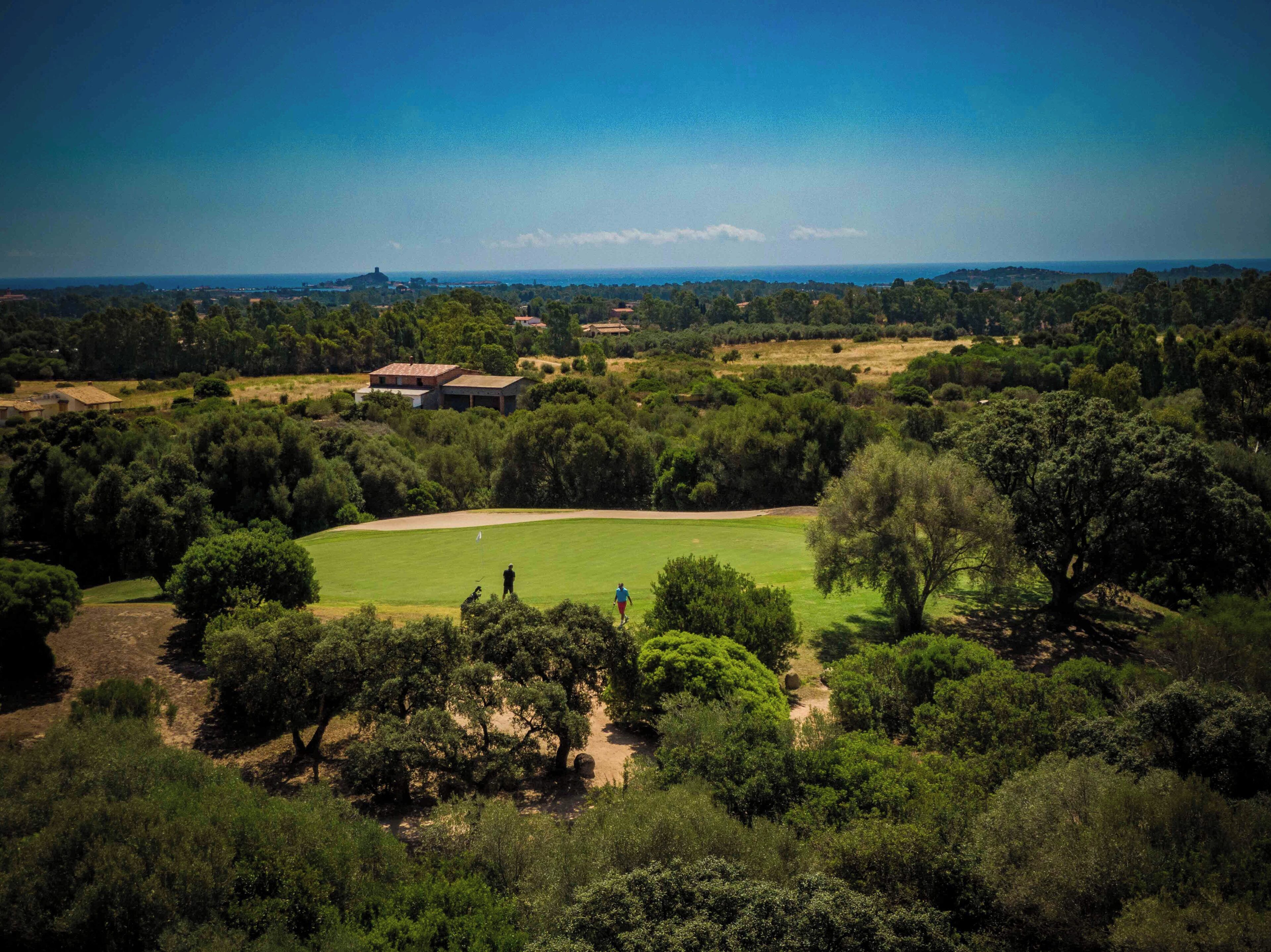 A golf course with a clubhouse, surrounded by dense trees and a clear sky.