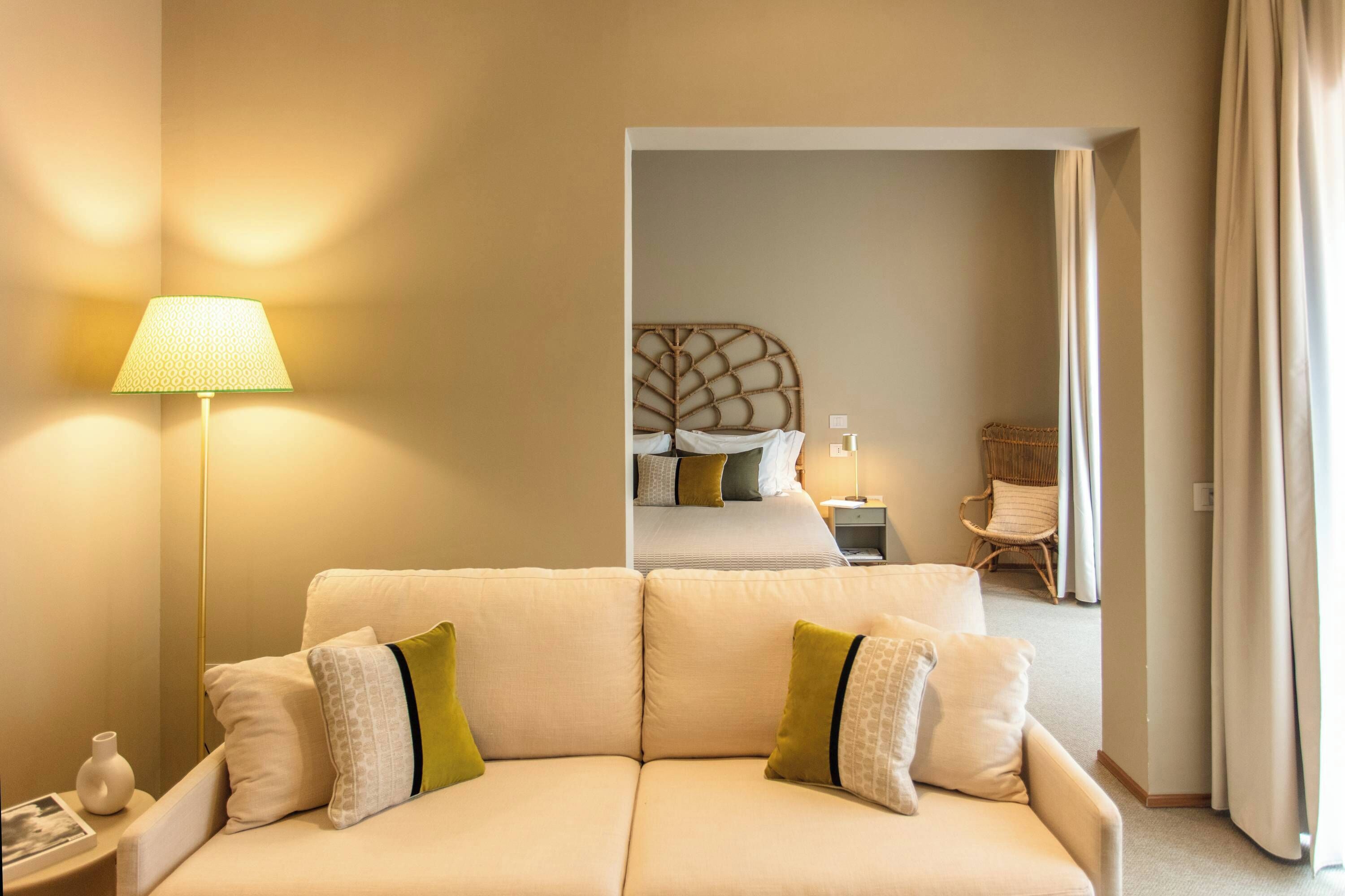 A hotel room with a sofa, a lamp, a bed with a leaf-patterned headboard, and a wicker chair.