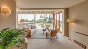Amplia sala de estar con sofá, sillas y una mesa de centro, ventanas grandes que ofrecen vistas al exterior y una planta en maceta.