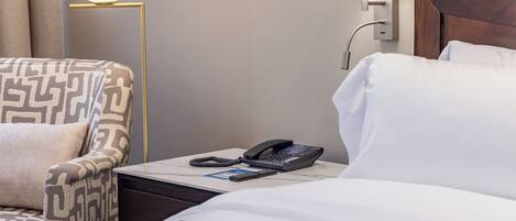 A hotel room with a patterned armchair, a bedside table with a phone and remote control, and a wall-mounted lamp.