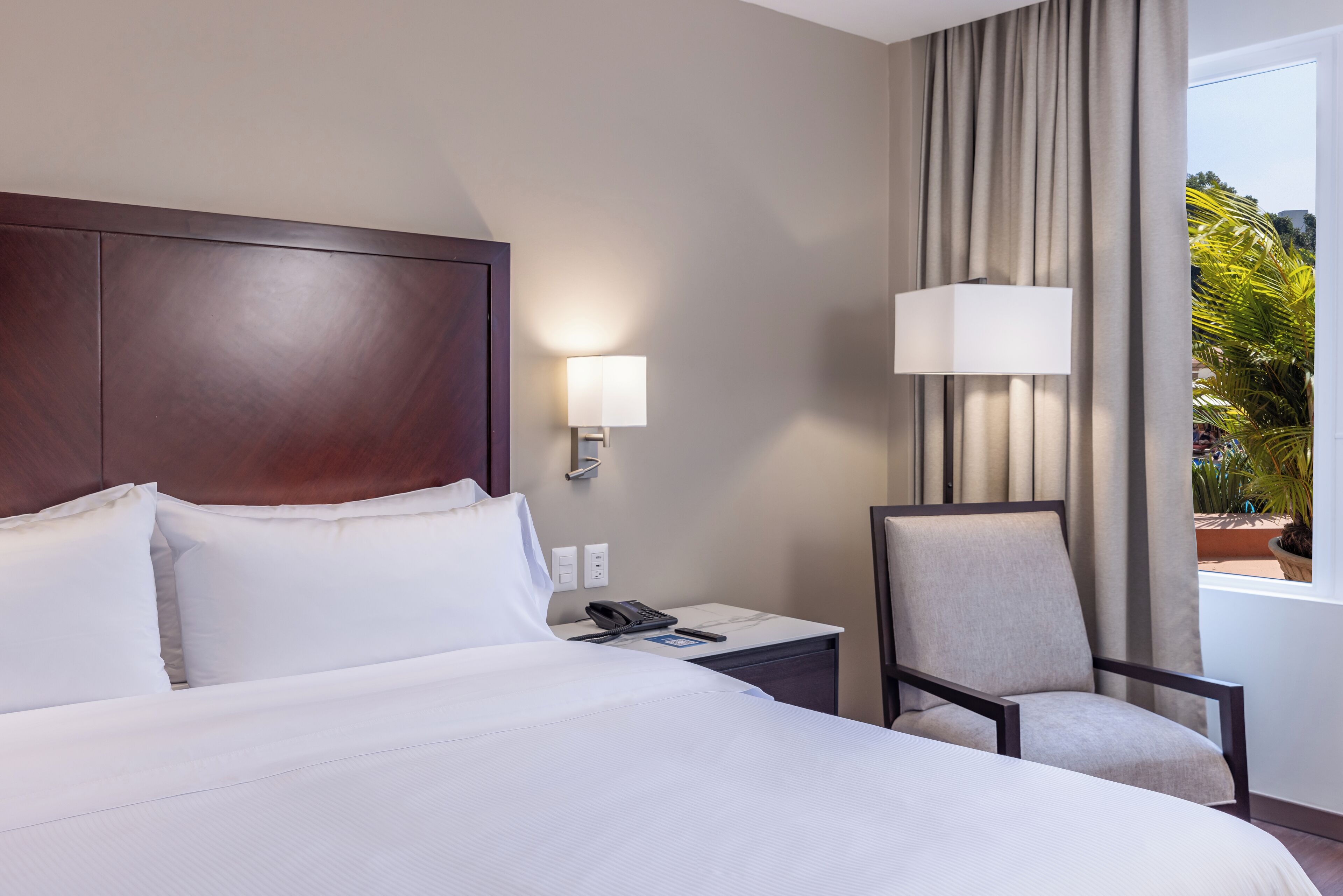 A hotel room with a large bed, a nightstand with a phone, a chair, and a window with curtains.