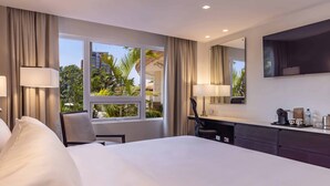 Une chambre d’hôtel avec un grand lit, un bureau avec une chaise, une télévision et une fenêtre donnant sur des palmiers et des immeubles.
