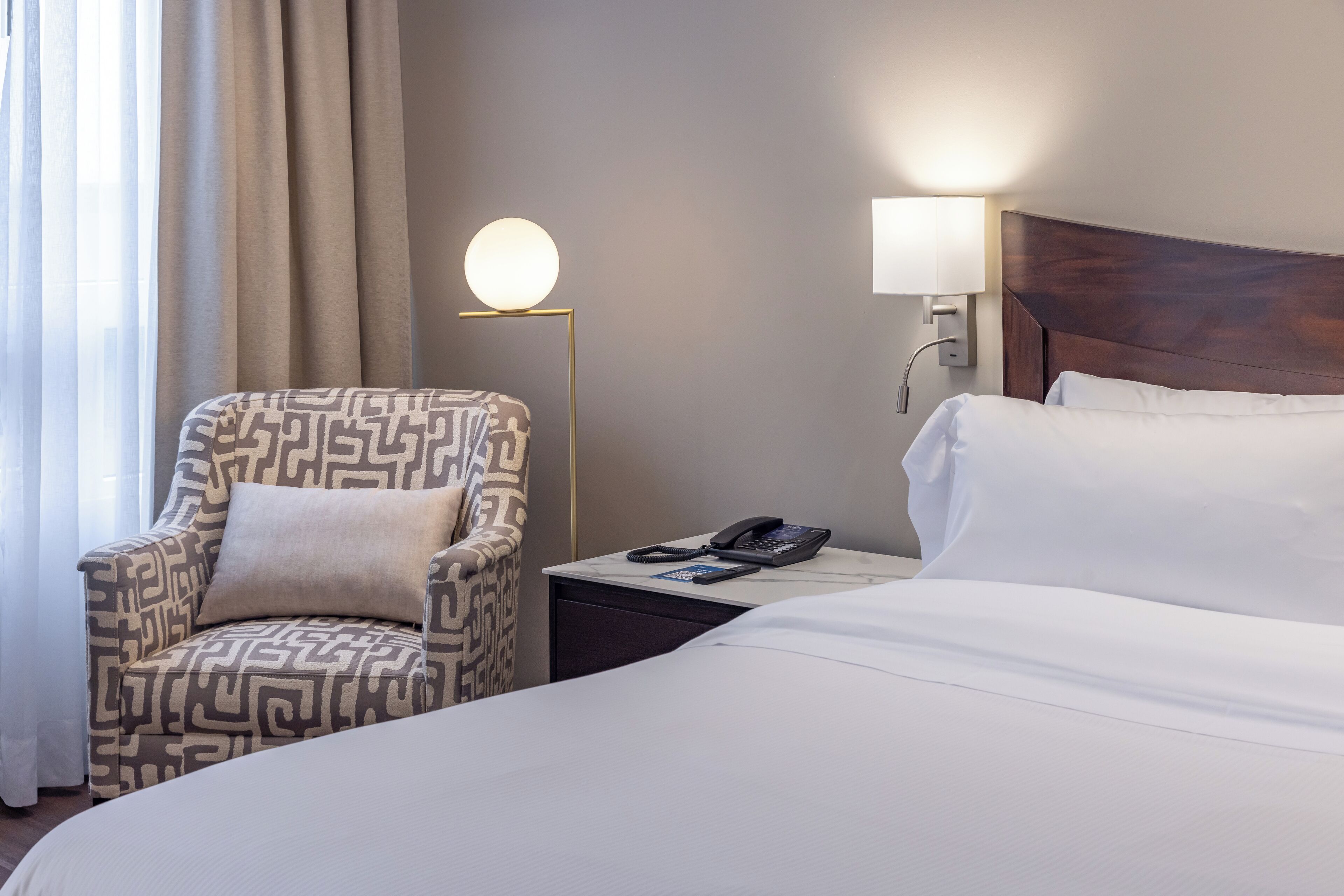 Une chambre d’hôtel avec un lit, un fauteuil à motifs, une table de chevet avec une lampe et un téléphone.