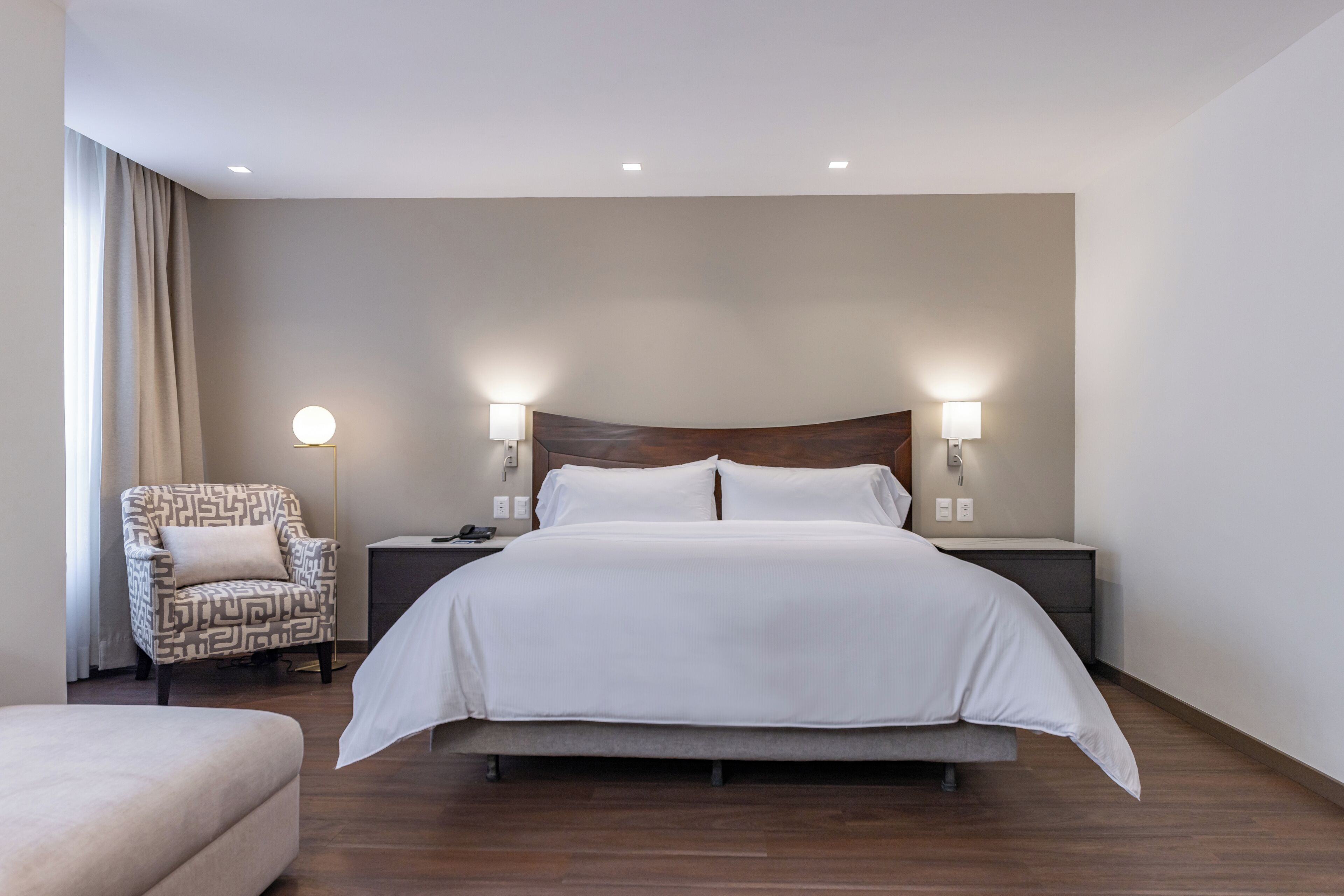 A modern hotel room with a large bed, two bedside tables, a patterned armchair, and a small sofa.