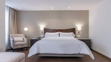 A modern hotel room with a large bed, two bedside tables, a patterned armchair, and a small sofa.