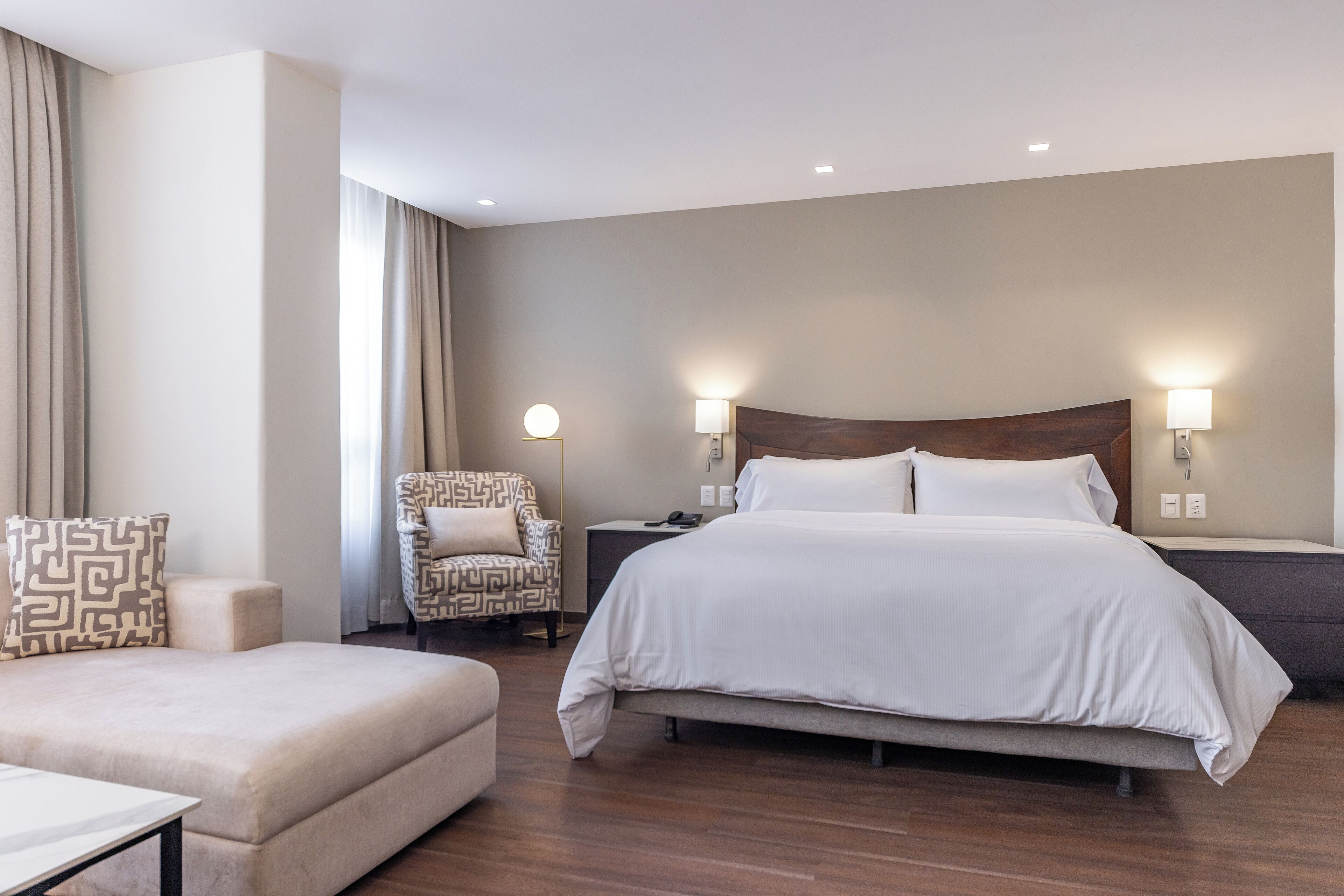 A hotel room with a bed, a chair, a nightstand, and a bedside table.