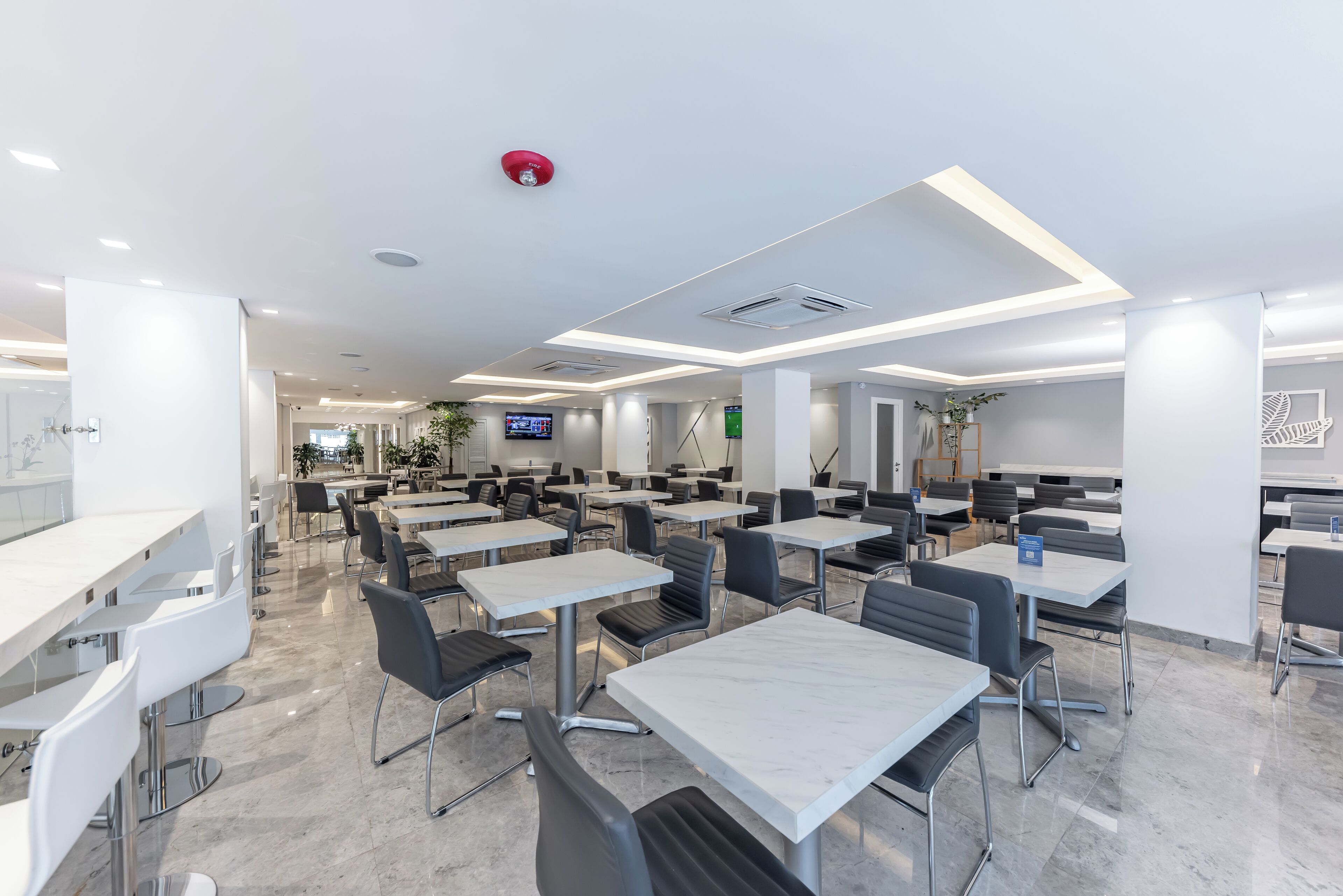 A modern dining area with white tables and black chairs, a flat-screen TV, and a red fire alarm on the ceiling.