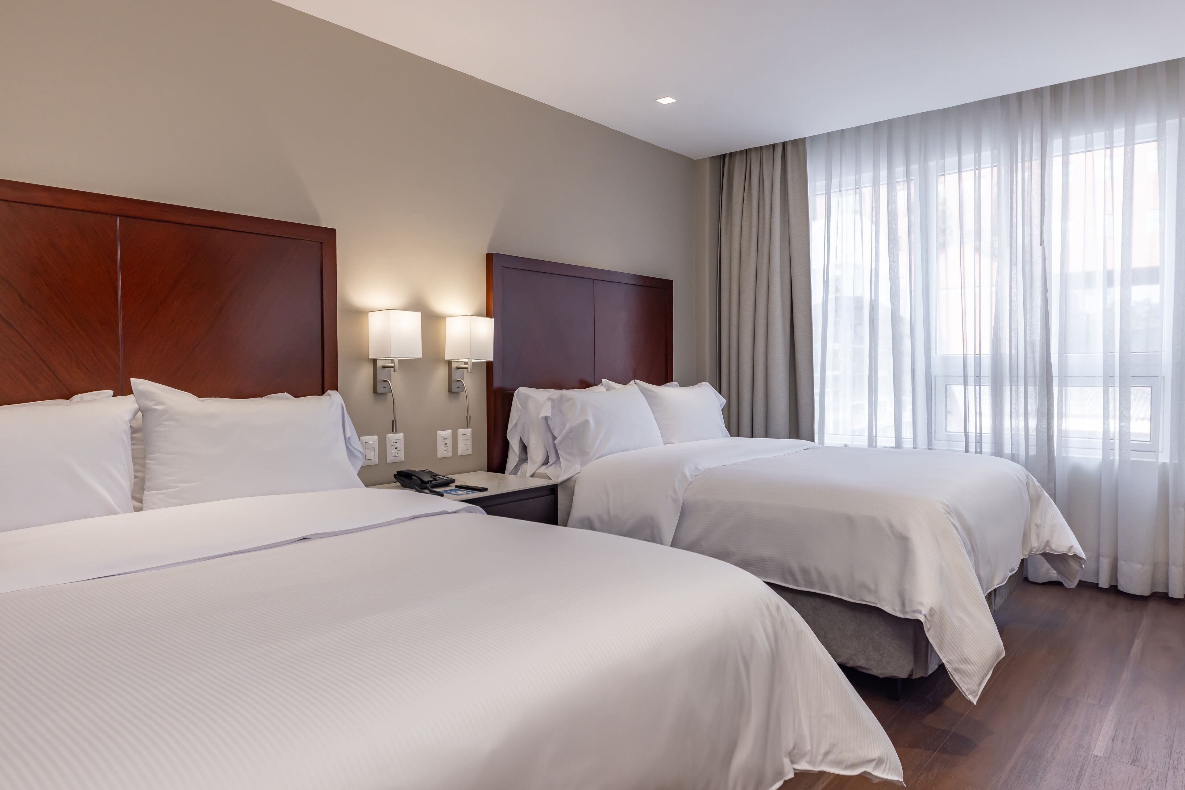 Habitación de hotel con dos camas, cabecero de madera, mesita de noche con lámpara y un ventanal con cortinas.