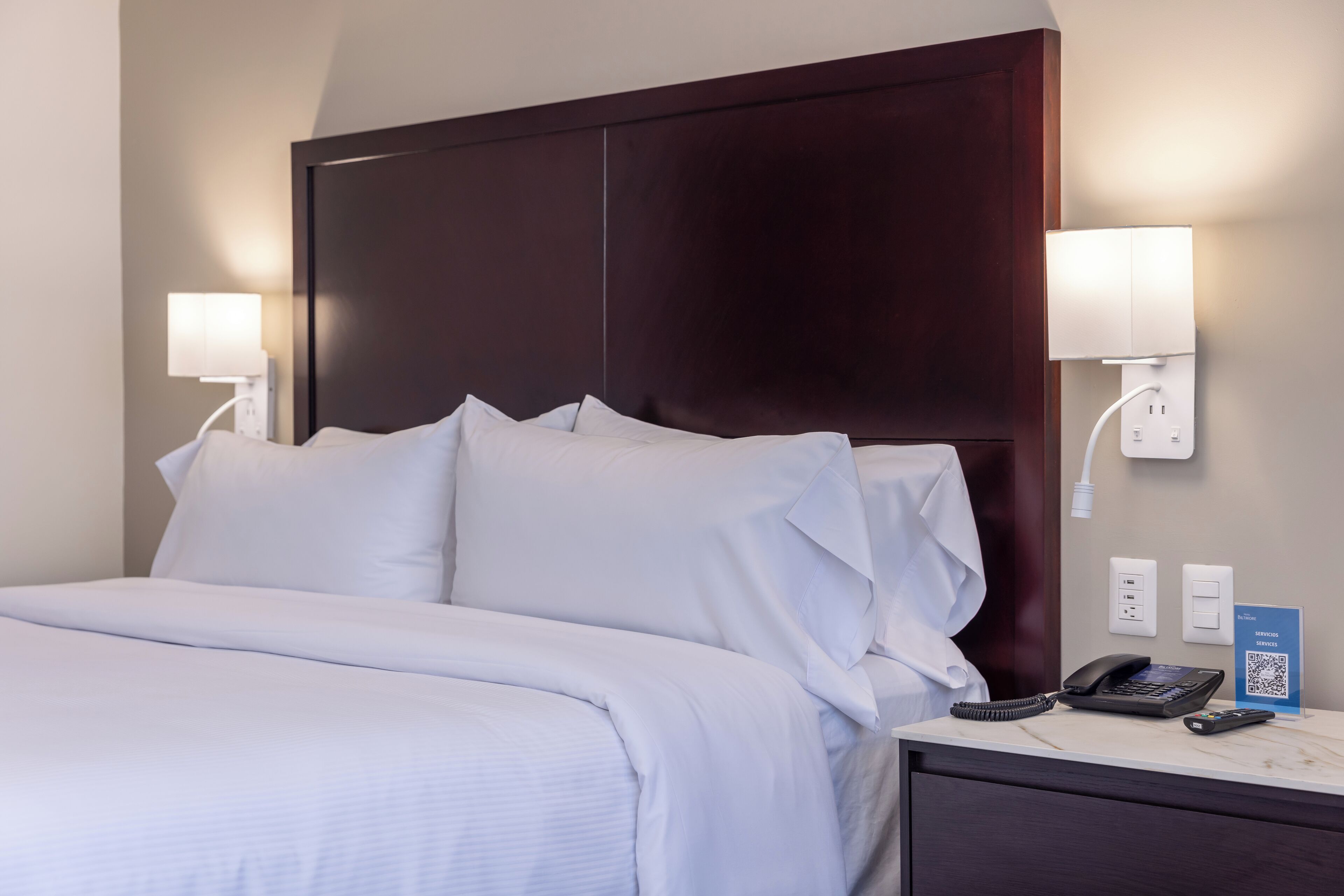Habitación de hotel con una cama grande, dos lámparas de noche, un teléfono y un código QR en la mesita de noche.