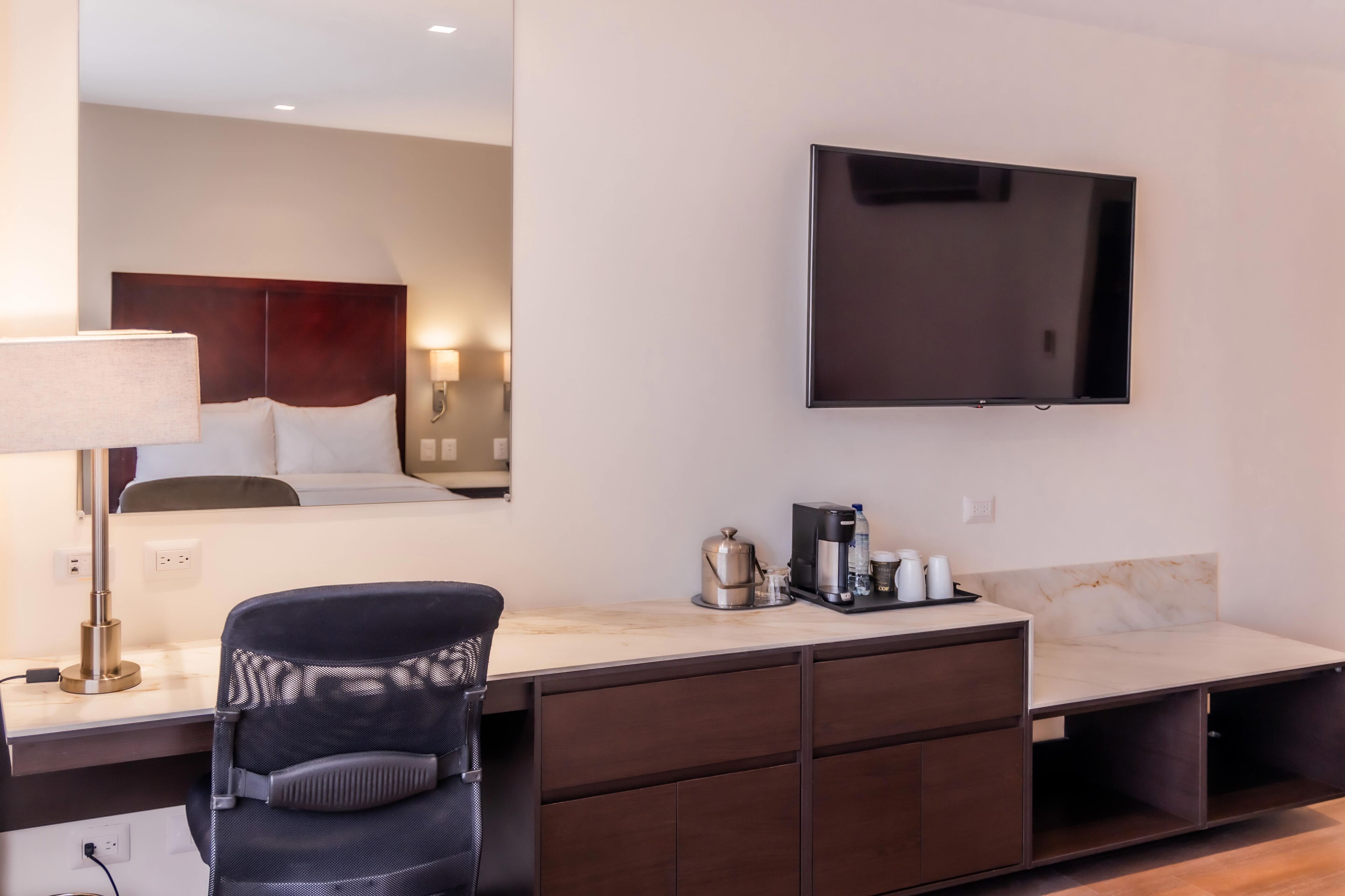 A hotel room with a bed, a desk with a chair, a flat-screen TV mounted on the wall, and a coffee maker on the desk.