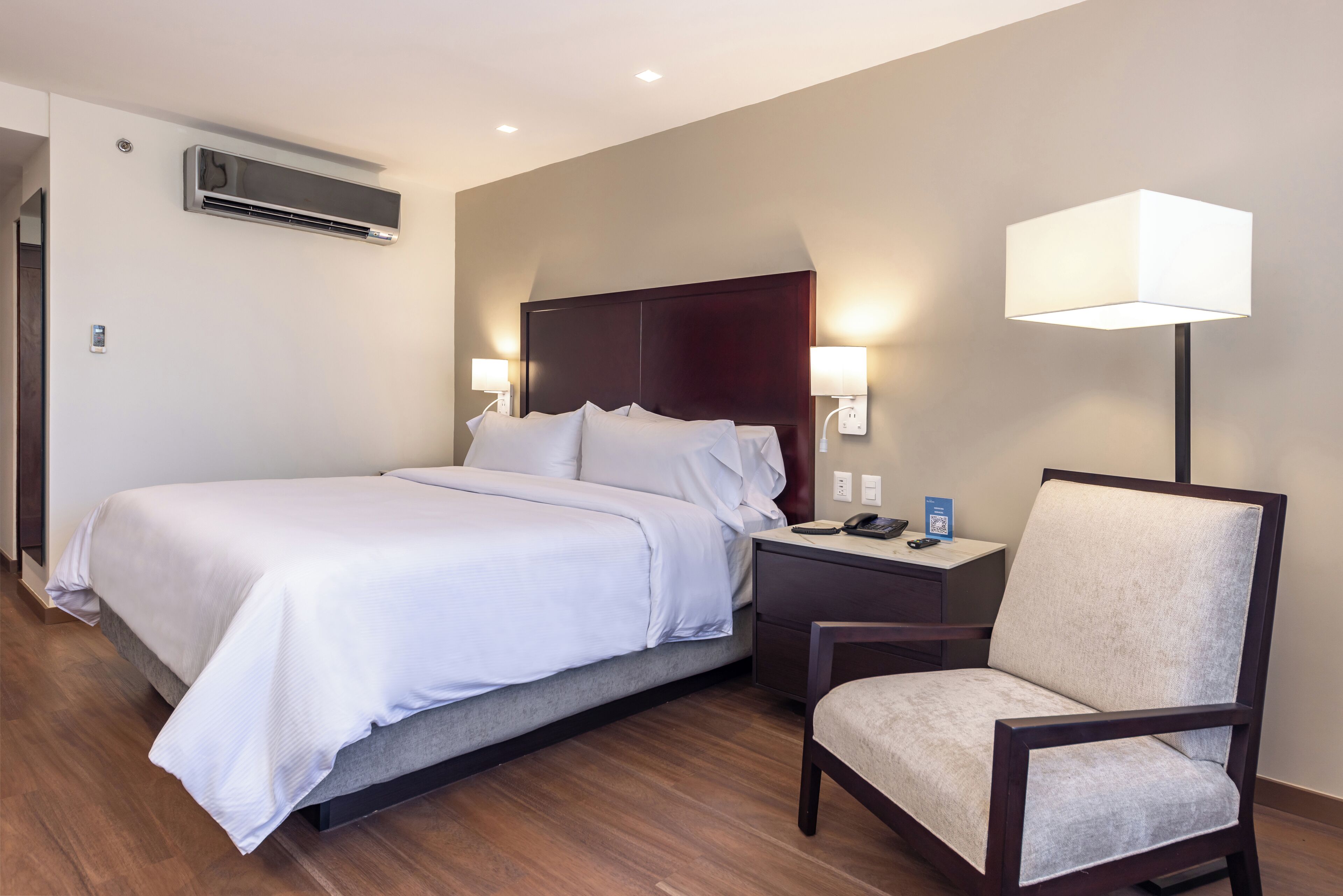 A hotel room with a bed, bedside table, chair, lamp, and air conditioning unit.