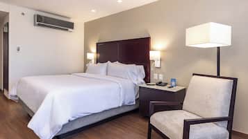 A hotel room with a bed, bedside table, chair, lamp, and air conditioning unit.