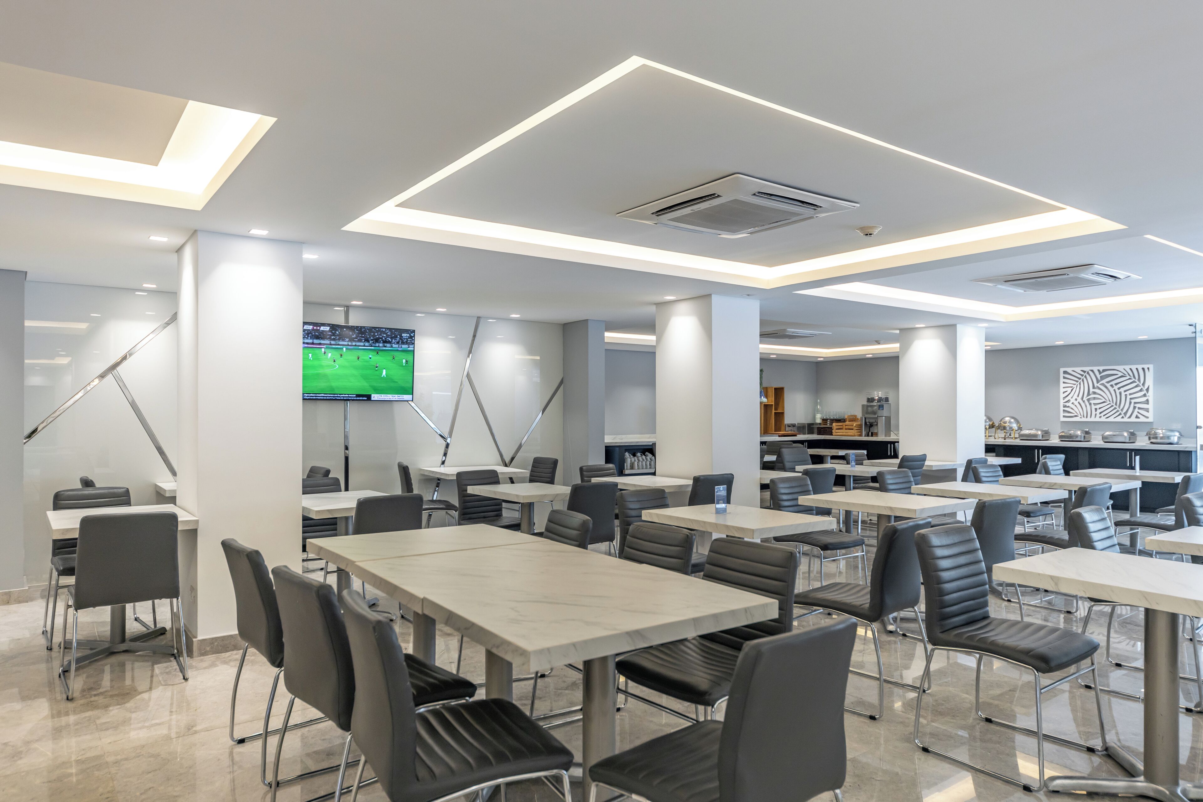 Un espace repas moderne avec plusieurs tables et chaises, un grand écran diffusant un événement sportif et, au fond, un coin cuisine.