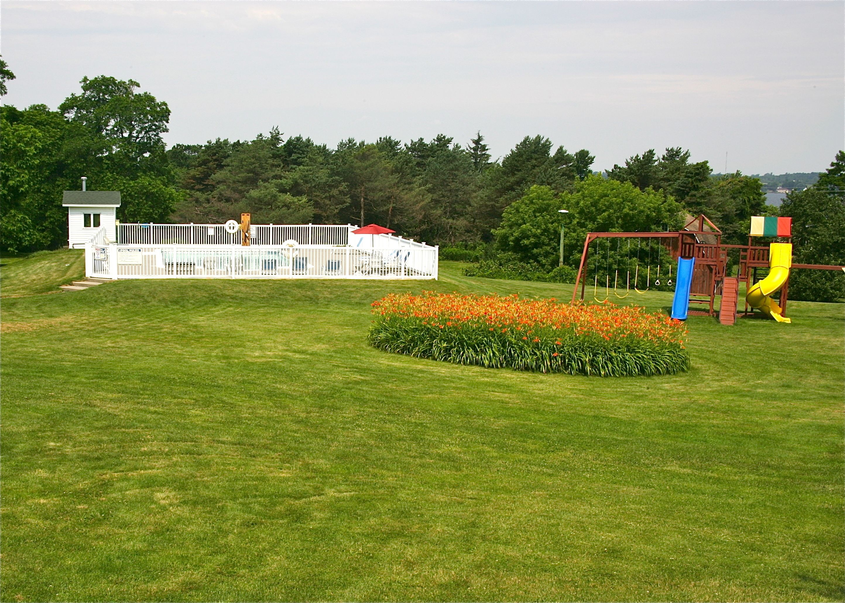 Un campo cubierto de césped con un parque infantil, un edificio blanco y un bosque al fondo.