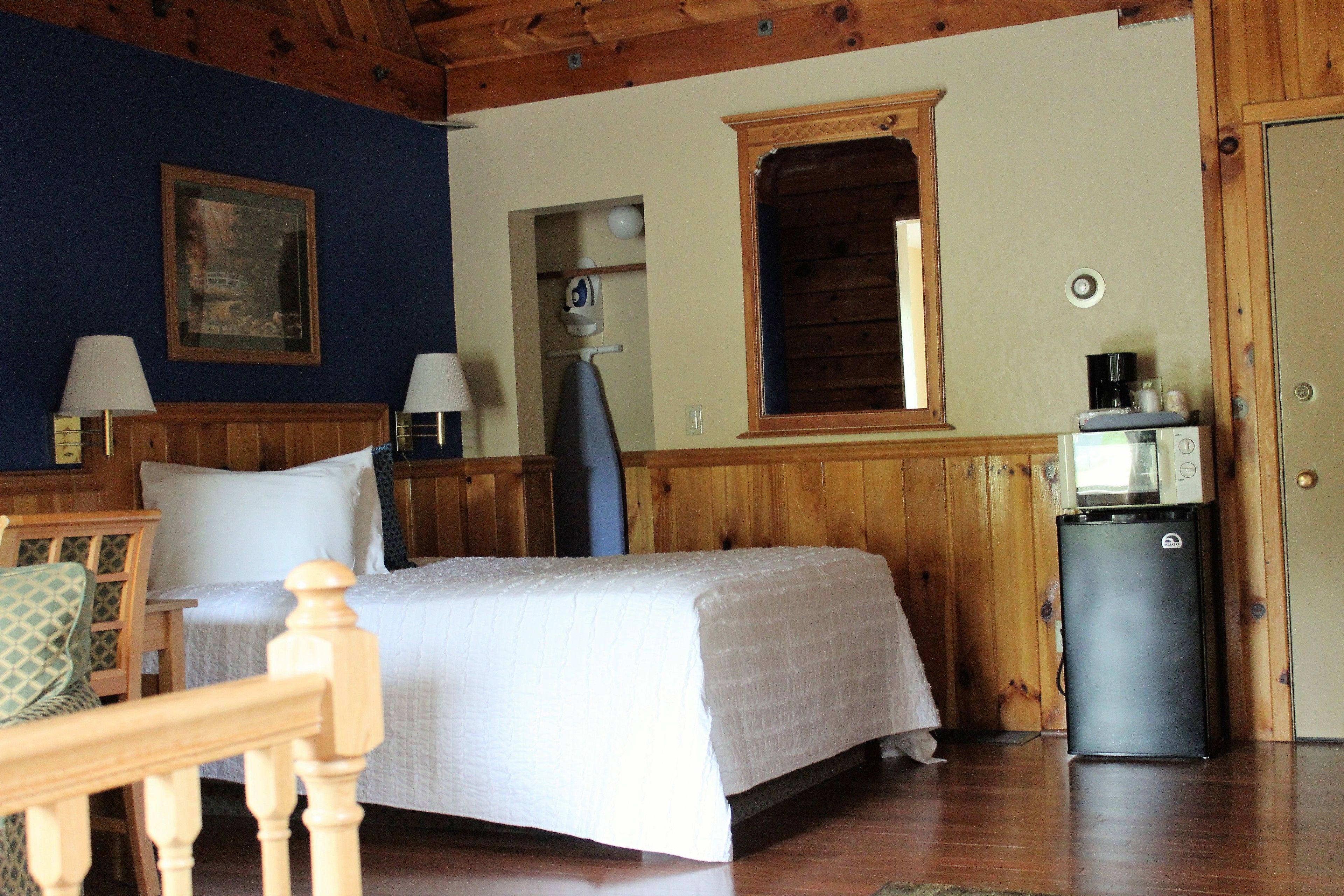 A room with a bed, a desk, a chair, a lamp, a refrigerator, and a painting on the wall.