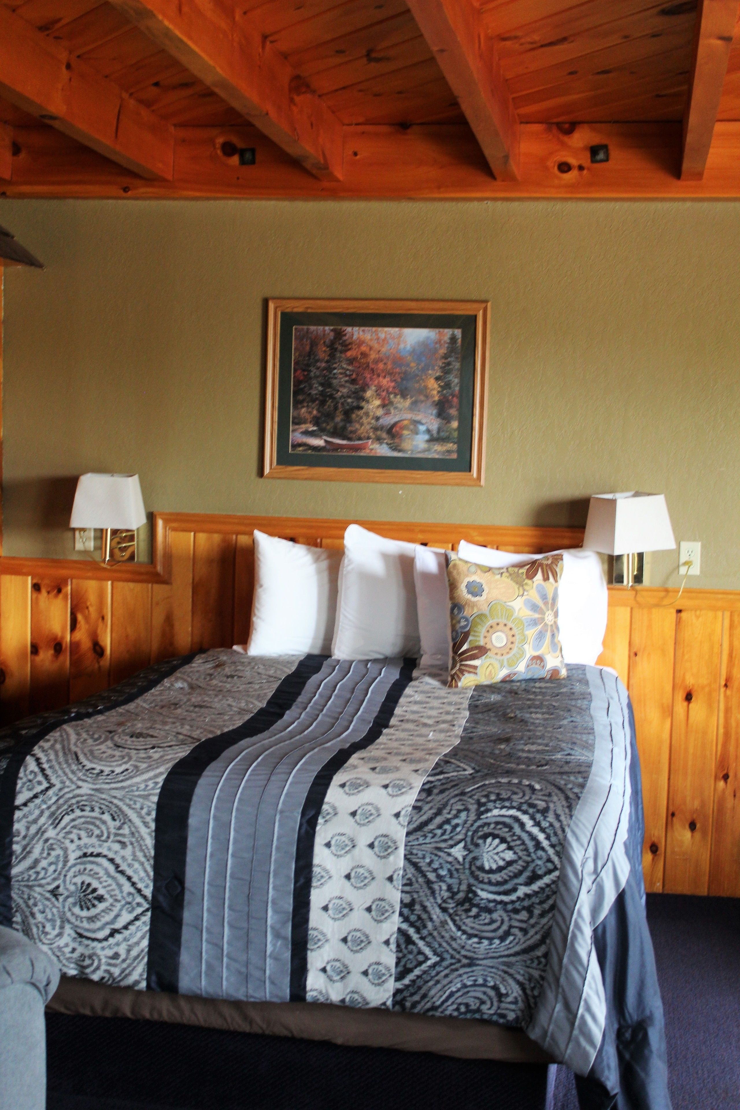 A wooden-framed bed with a patterned bedspread, two bedside lamps, and a framed painting on the wall.