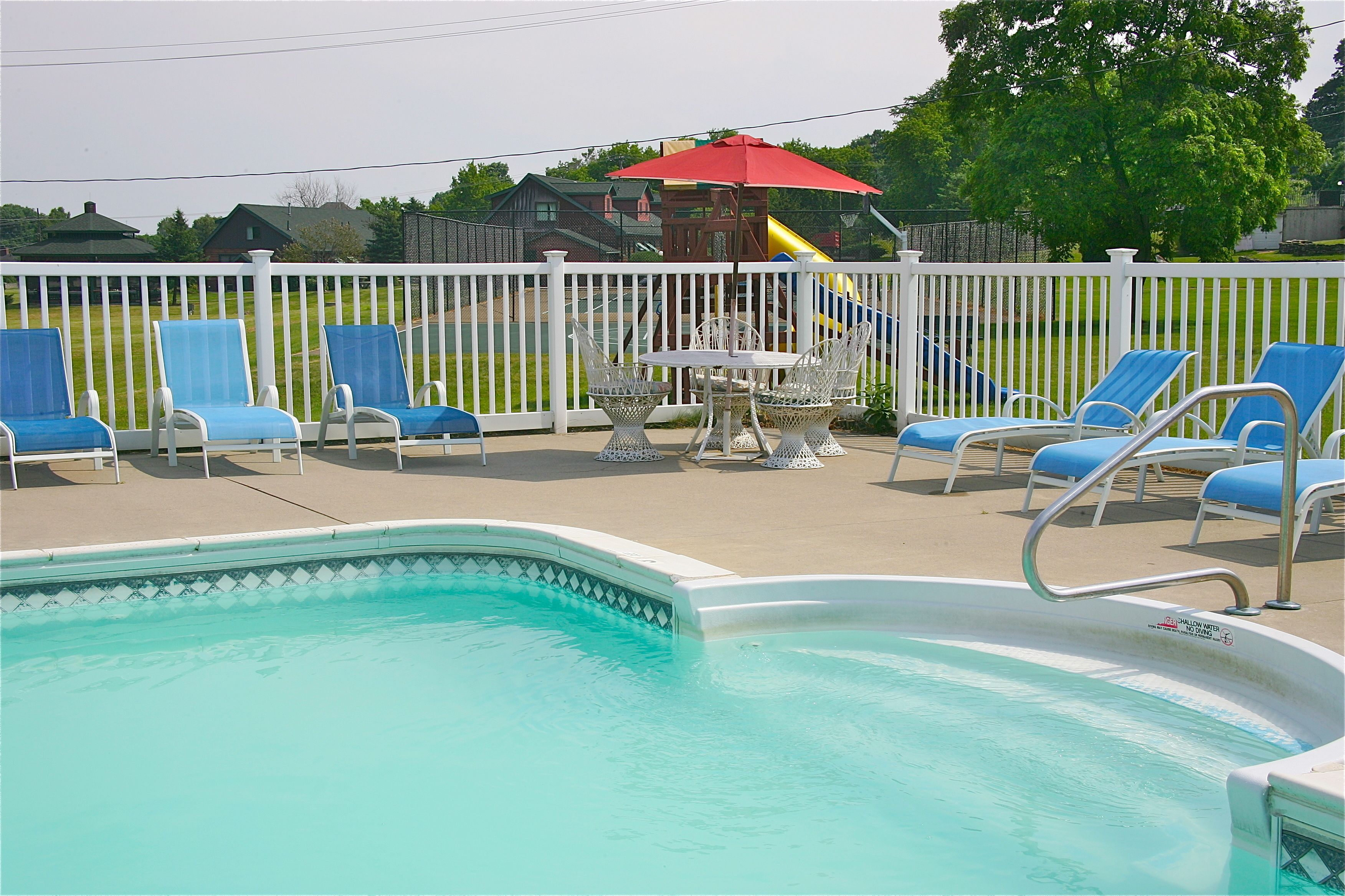 A swimming pool with blue lounge chairs and a table with chairs, surrounded by a white fence.