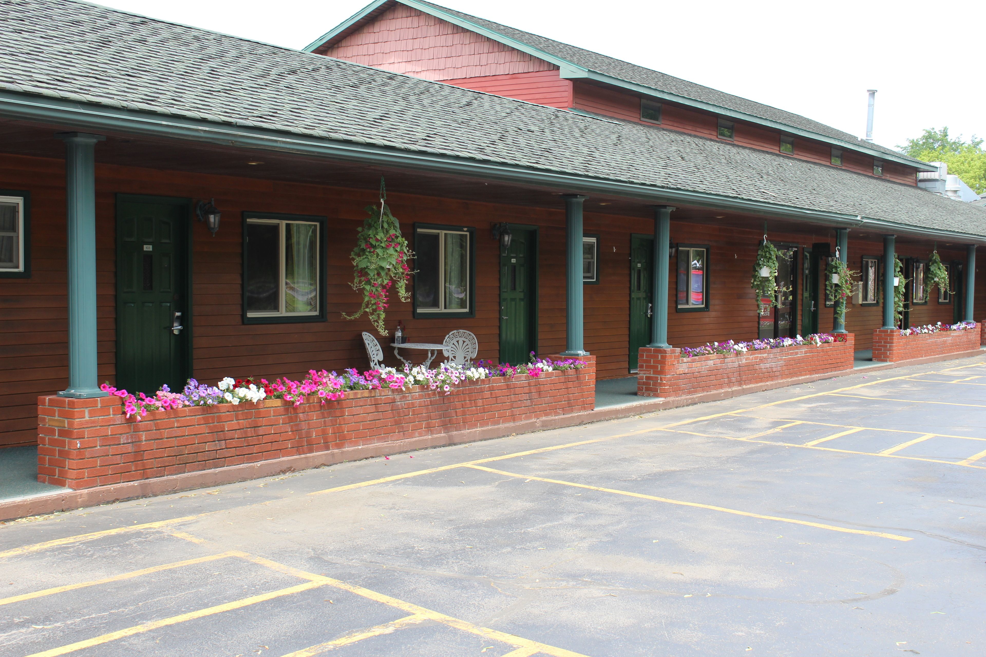 A row of wooden buildings with green doors and windows, a brick planter with flowers, and a parking lot.
