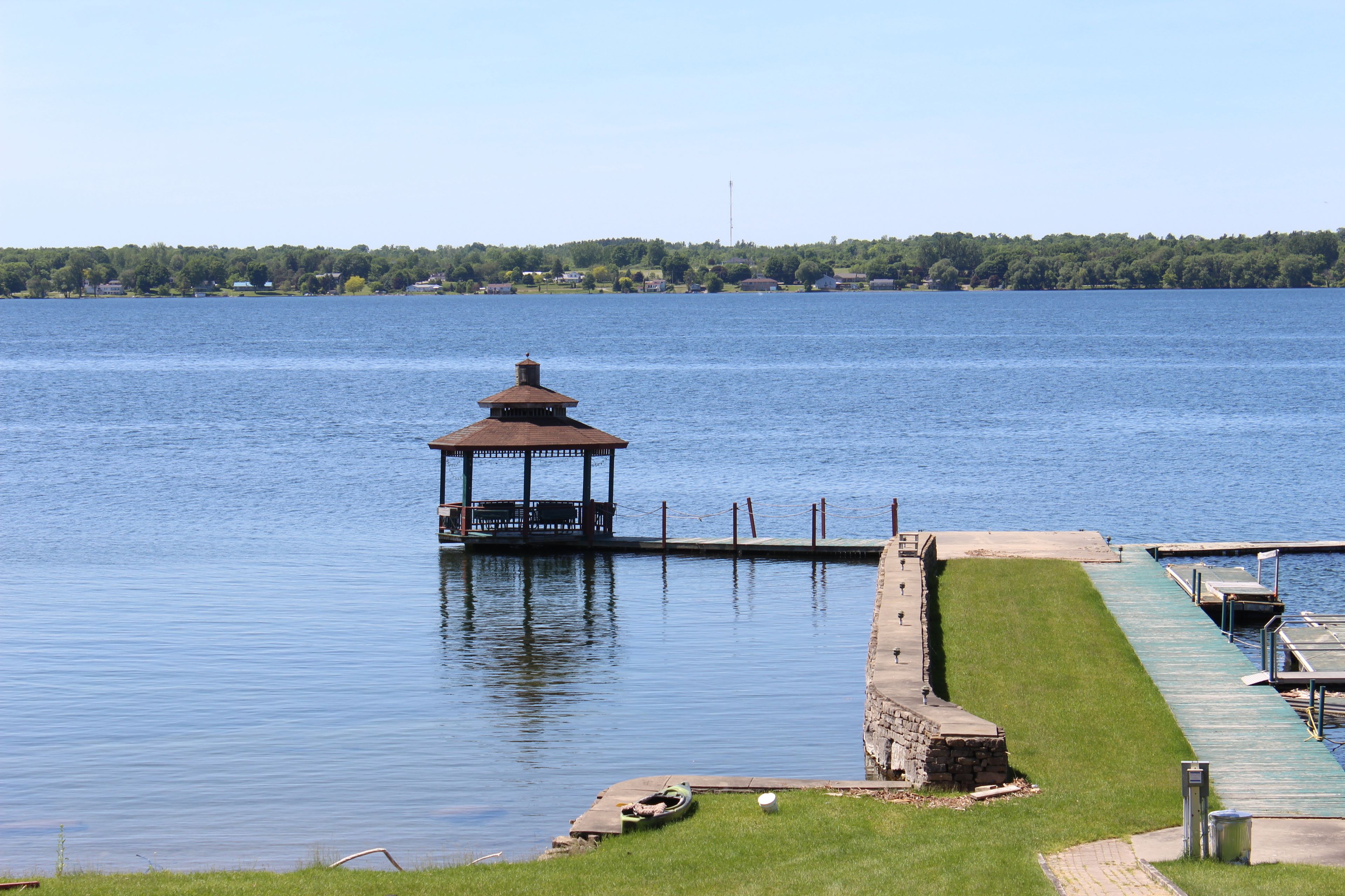 A gazebo on a dock extending into a calm lake, surrounded by a grassy area and a stone wall.