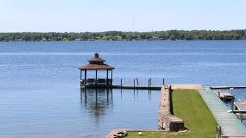 A gazebo on a dock extending into a calm lake, surrounded by a grassy area and a stone wall.