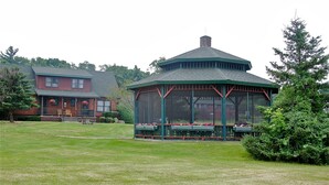 A gazebo with a green roof and wooden structure, surrounded by a grassy lawn and a red building in the background. - The Stonefence Resort (Ogdensburg)