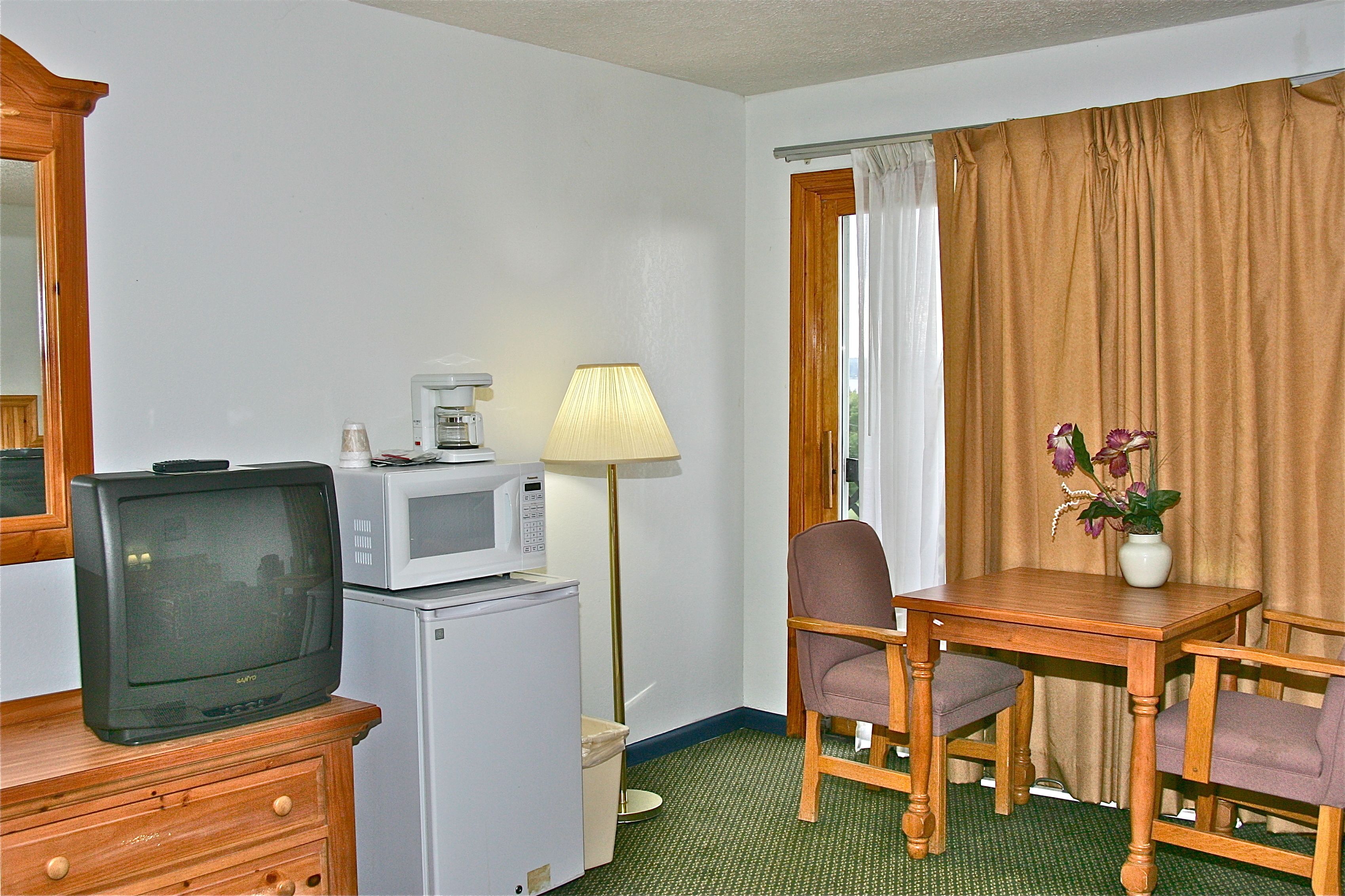 Habitación de hotel compacta con televisión, microondas, refrigerador pequeño, mesa de madera, dos sillas, lámpara y un florero con flores.