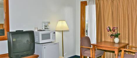 Habitación de hotel compacta con televisión, microondas, refrigerador pequeño, mesa de madera, dos sillas, lámpara y un florero con flores.