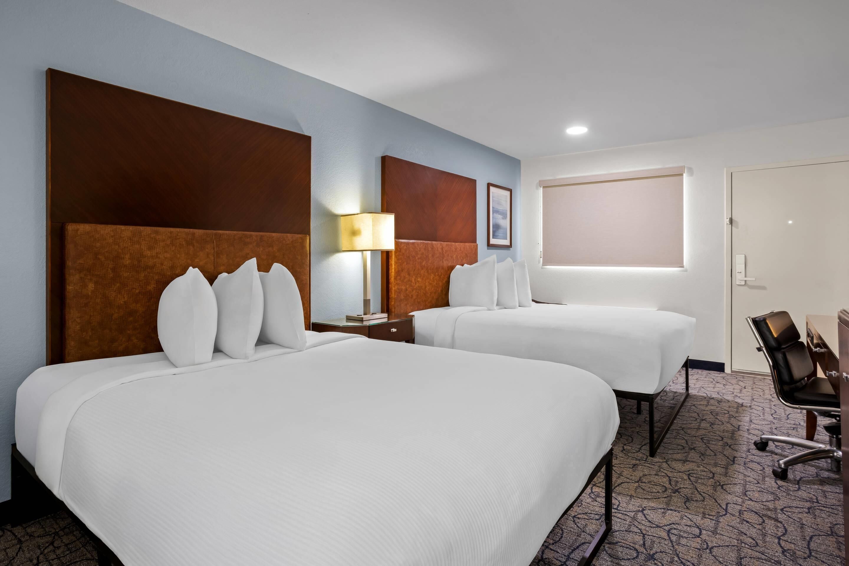 A hotel room with two beds, a desk, a chair, a lamp, and a window.