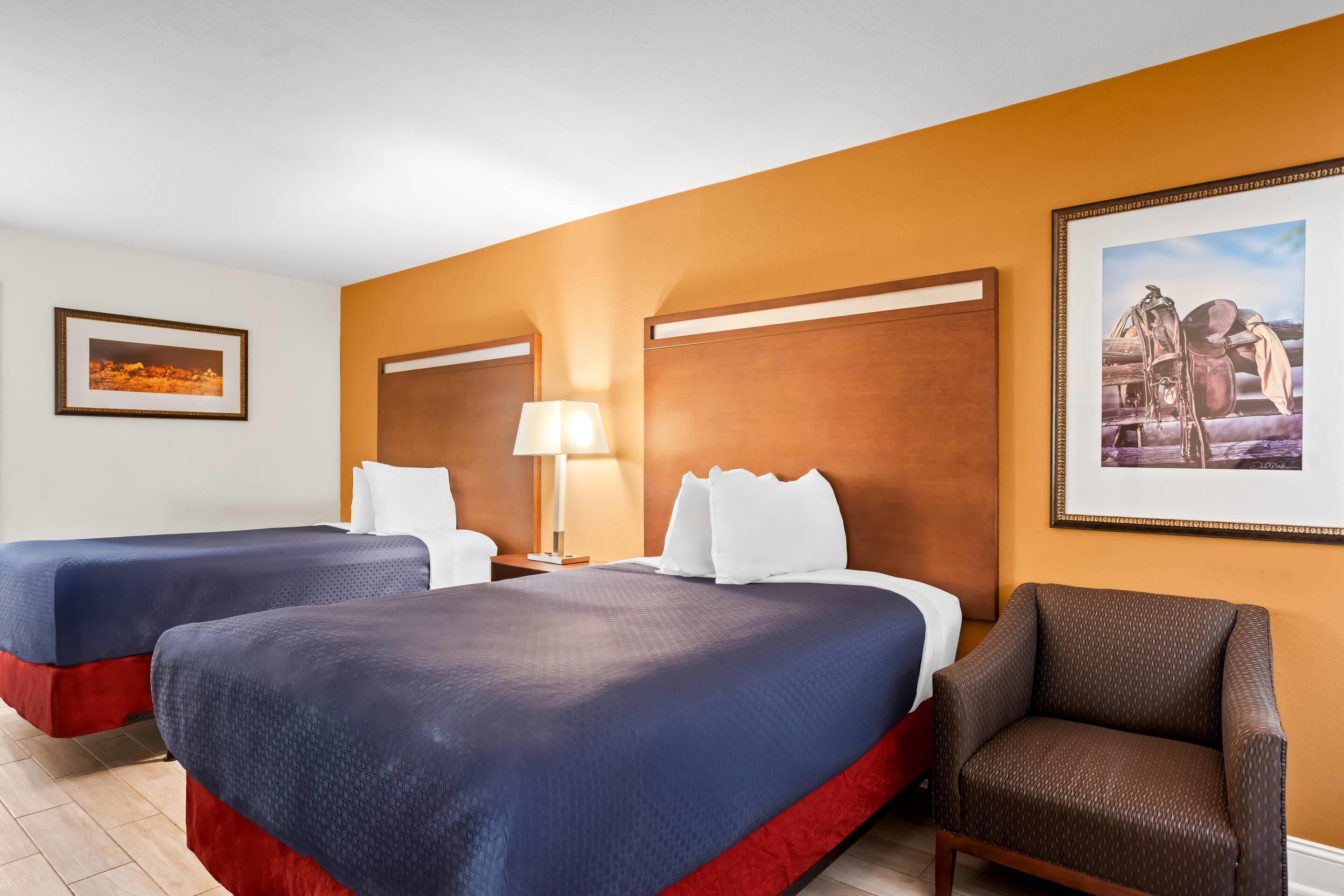 A hotel room with two beds, a nightstand with a lamp, a chair, and two framed pictures on the wall.