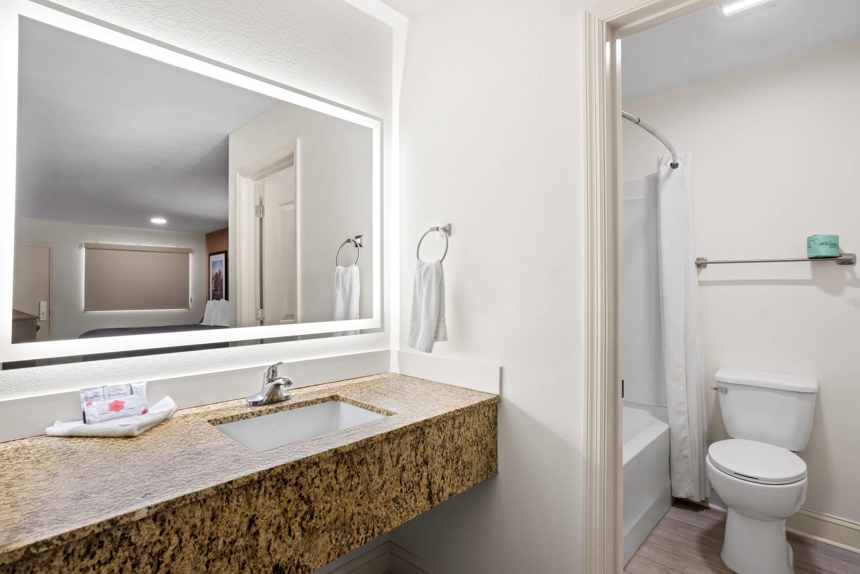 Un baño con lavamanos de encimera de granito, un espejo grande, inodoro y zona de ducha.