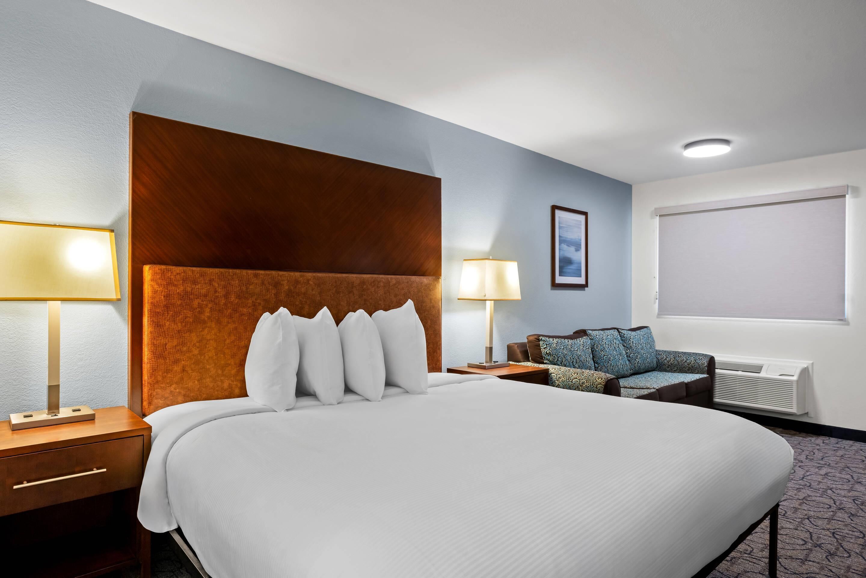 A hotel room with a large bed, two bedside tables, a sofa, a window with blinds, and a wall-mounted air conditioner.