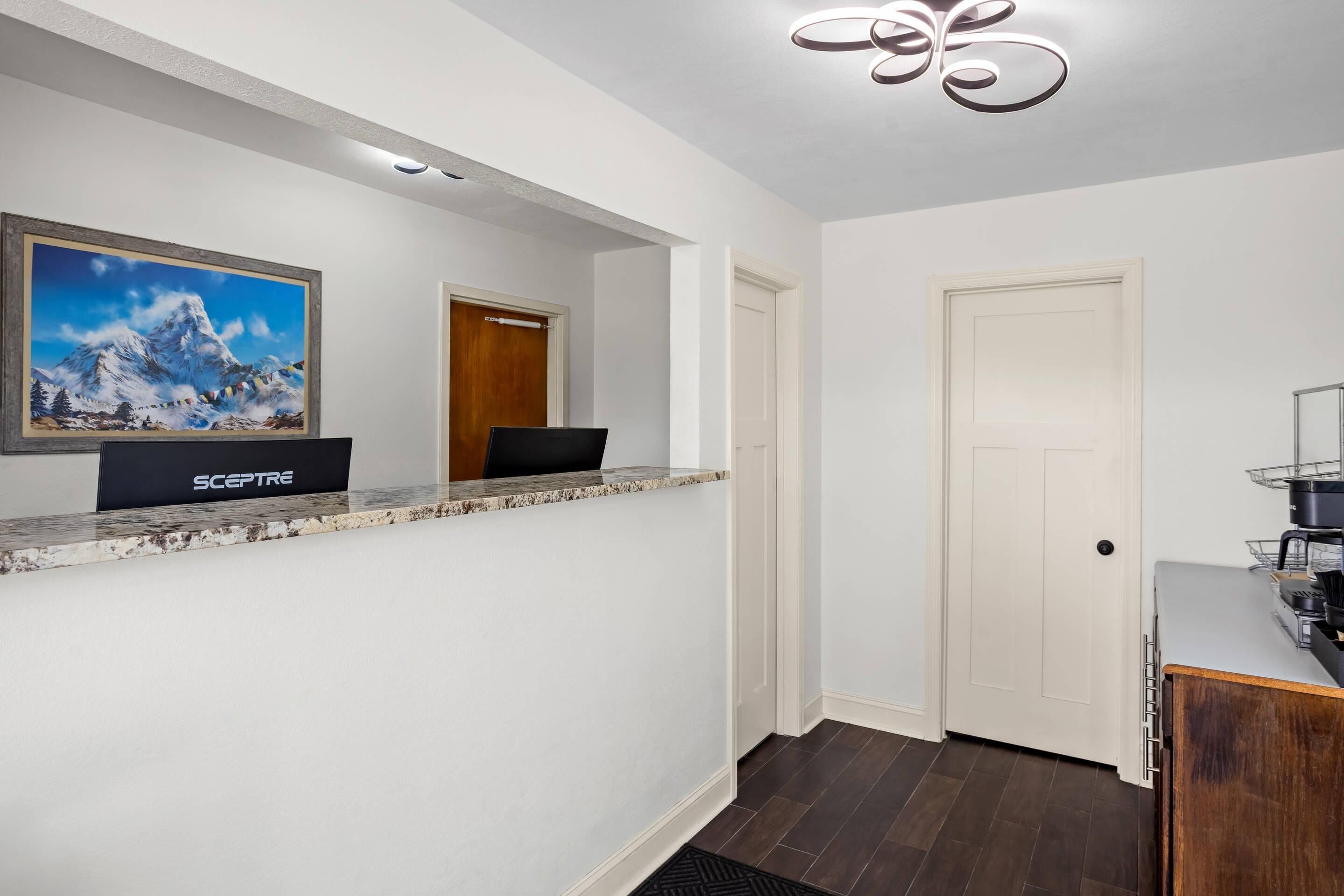 A hotel reception area with a reception desk, a framed painting of a mountain landscape, a door, and a coffee machine.